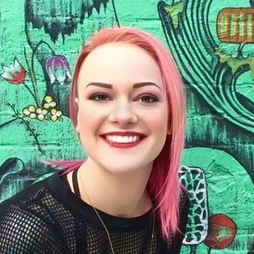 Smiling young woman with pink hair standing in front of a colorful, graffiti-style mural.