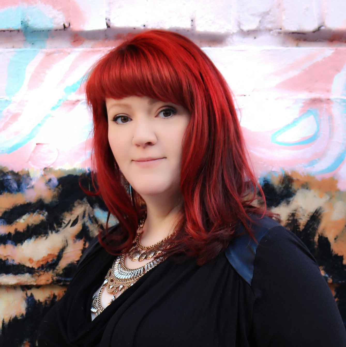 A woman with red hair taking a selfie in front of a colorful graffiti wall. She is wearing a black top and multiple layered necklaces.