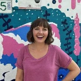 Smiling woman with shoulder-length brown hair and bangs, wearing a pink t-shirt, standing in front of a colorful, abstract mural with pink, blue, and teal shapes and patterns.