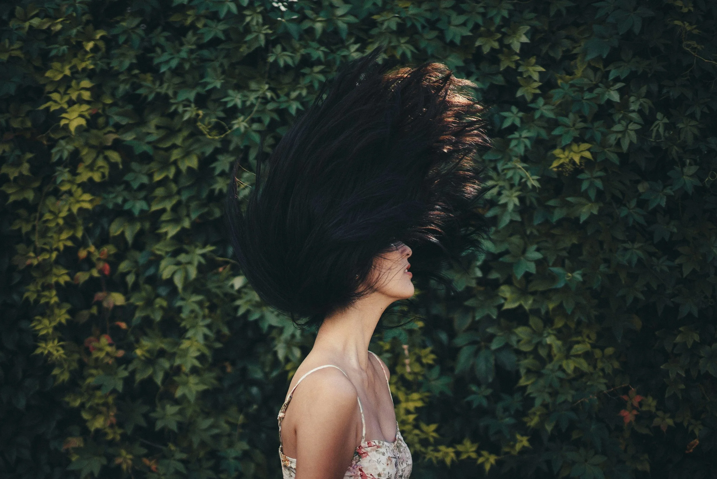 A woman with dark hair wearing a floral dress with thin straps, standing in front of a green leafy wall, with her hair blown upward