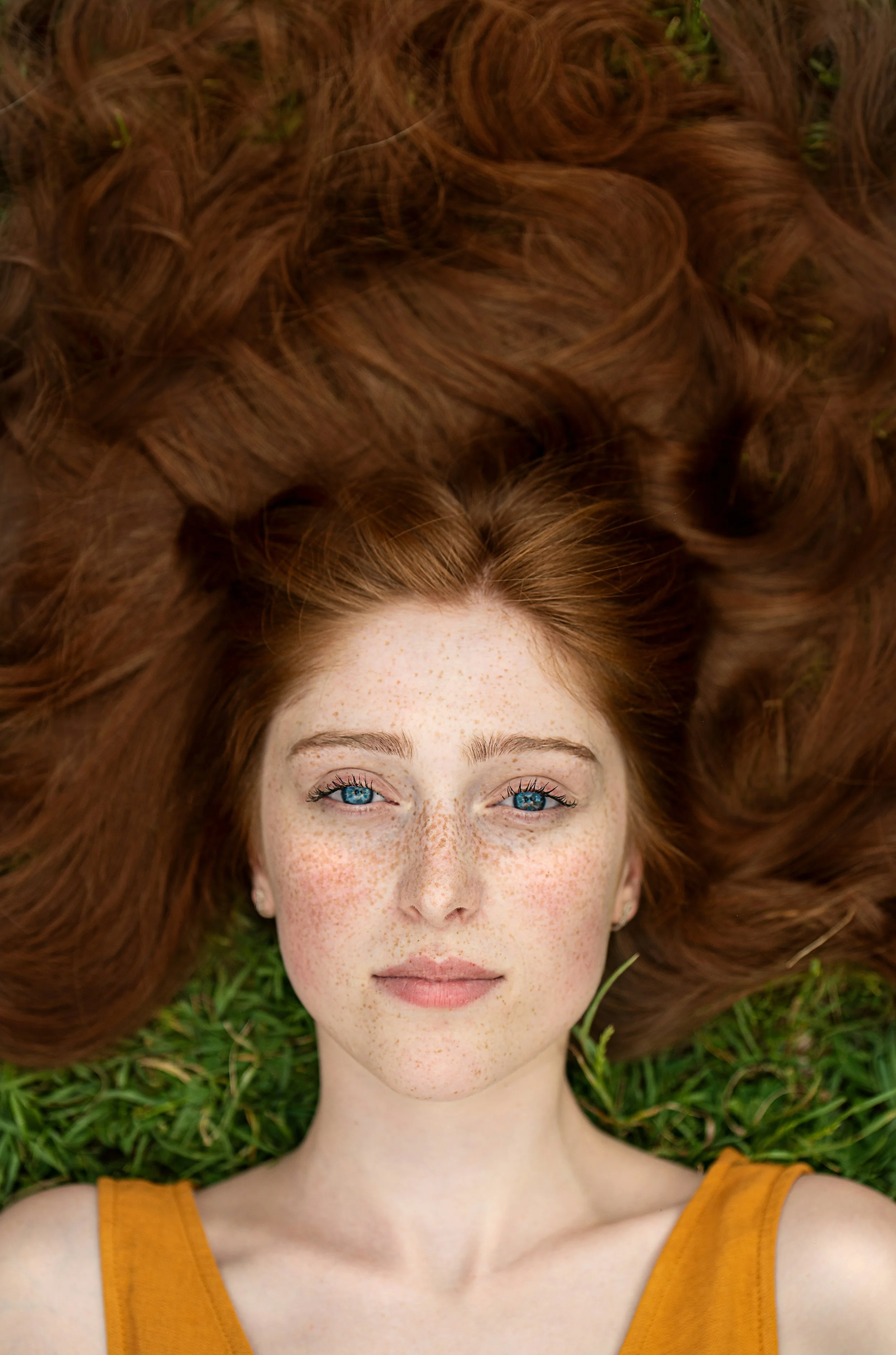 A woman with red hair and blue eyes lying on grass, wearing an orange tank top.