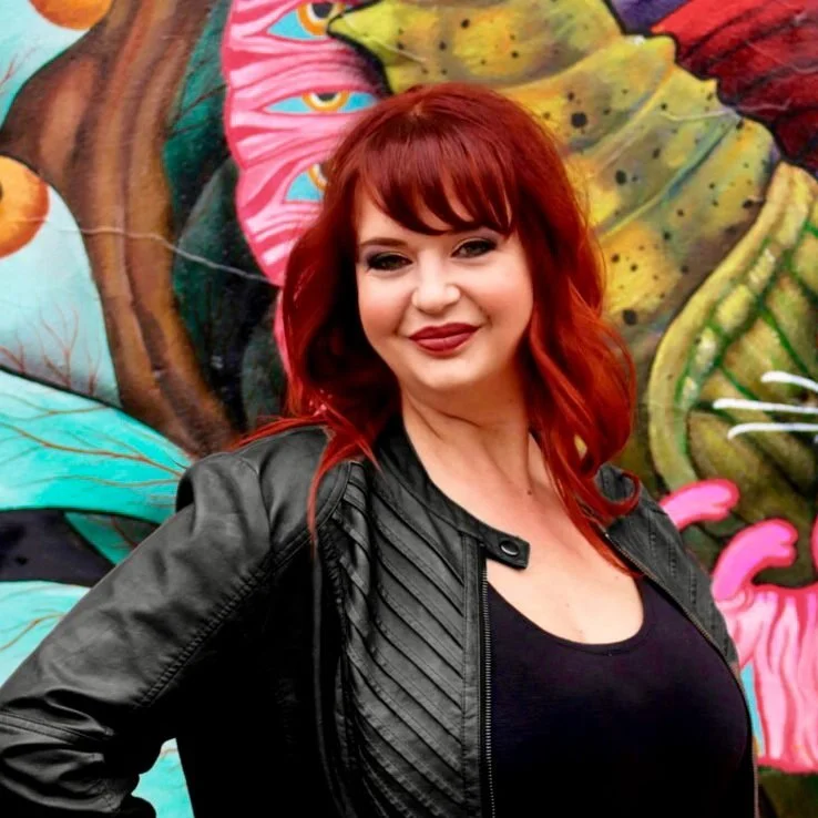 A woman with red hair smiling in front of a colorful mural.
