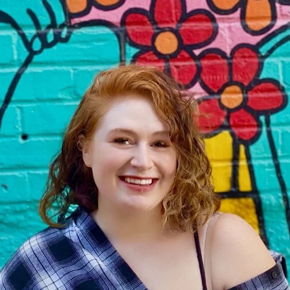 A young woman with curly, red hair smiling in front of a colorful mural with flowers and abstract designs.