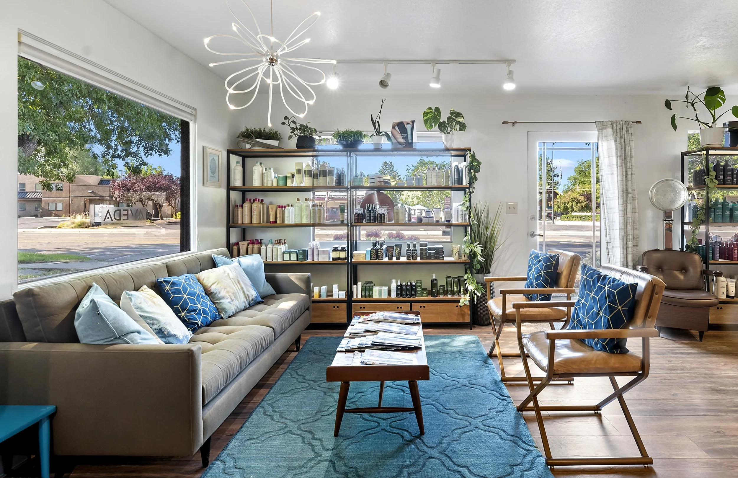 A cozy salon interior with a beige tufted sofa adorned with blue and beige pillows, a wooden coffee table, and wooden chairs with blue patterned pillows. Large windows show trees outside, and a glass shelf displays skincare and haircare products. Modern lighting fixtures hang from the ceiling, and there are several houseplants and curtains in the space.