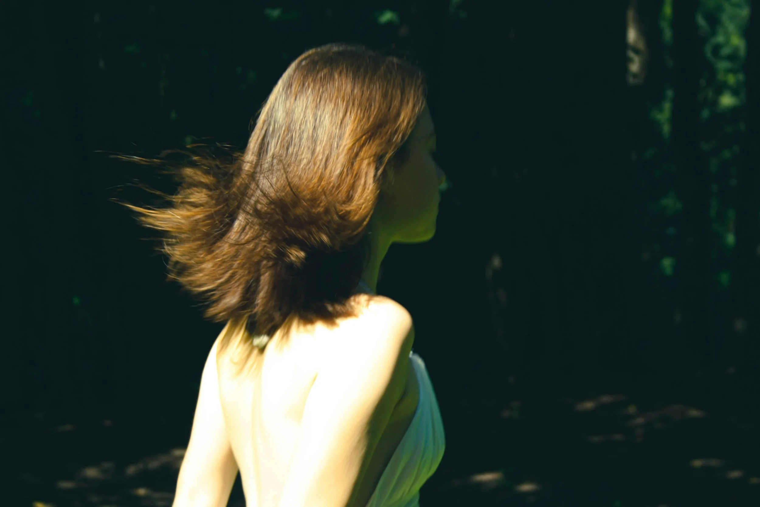 A woman with shoulder-length red hair, facing away, standing outdoors in a wooded area with dappled sunlight.