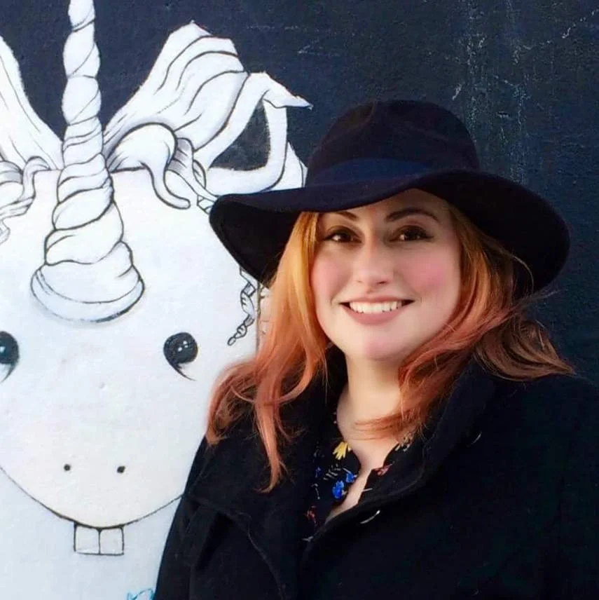A woman with orange hair and a black hat smiling in front of a black and white unicorn mural.