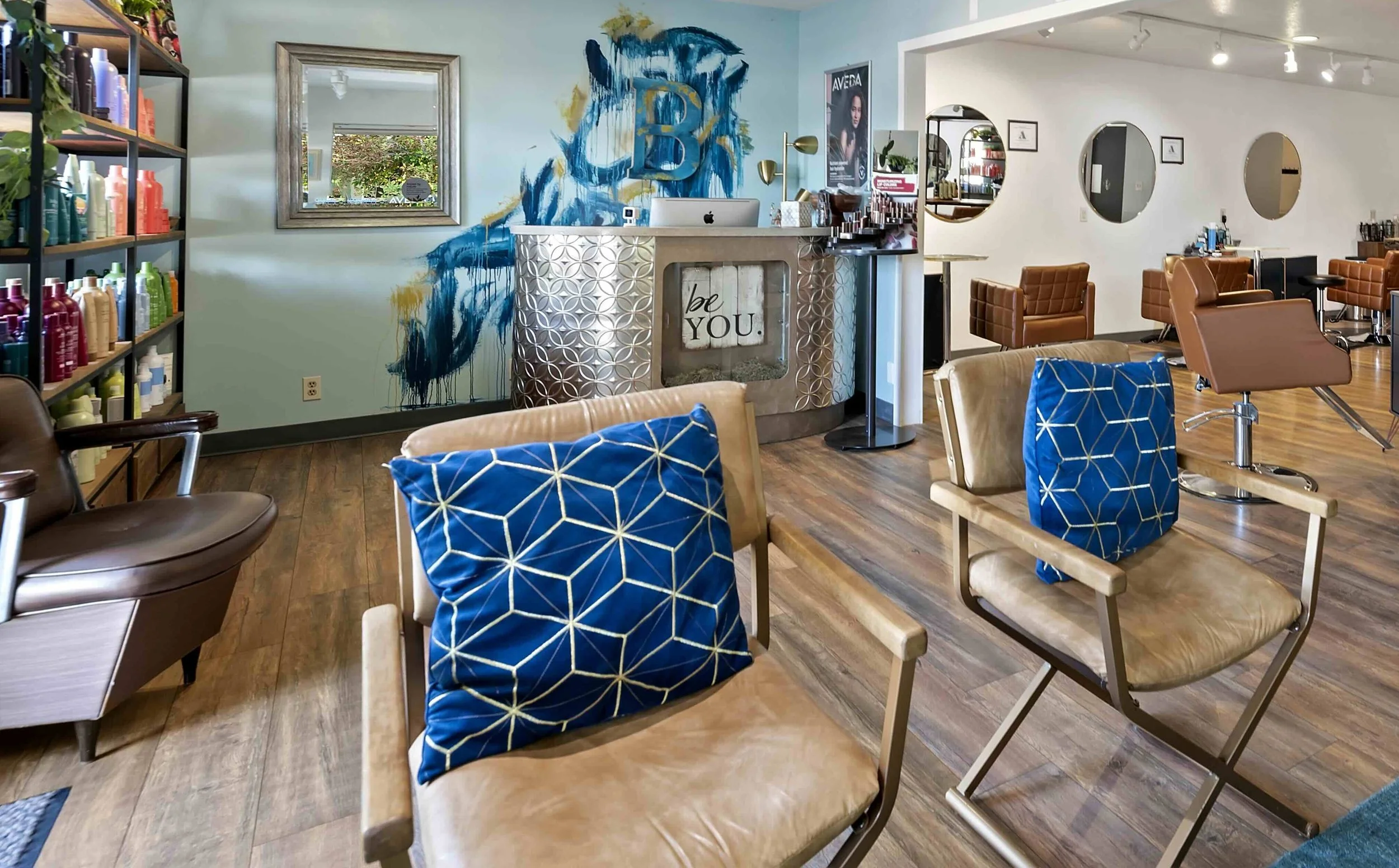 Interior of a modern salon with brown styling chairs, a reception desk with a sign that says "be YOU," colorful wall art, shelves with hair products, large mirrors, and wooden flooring.
