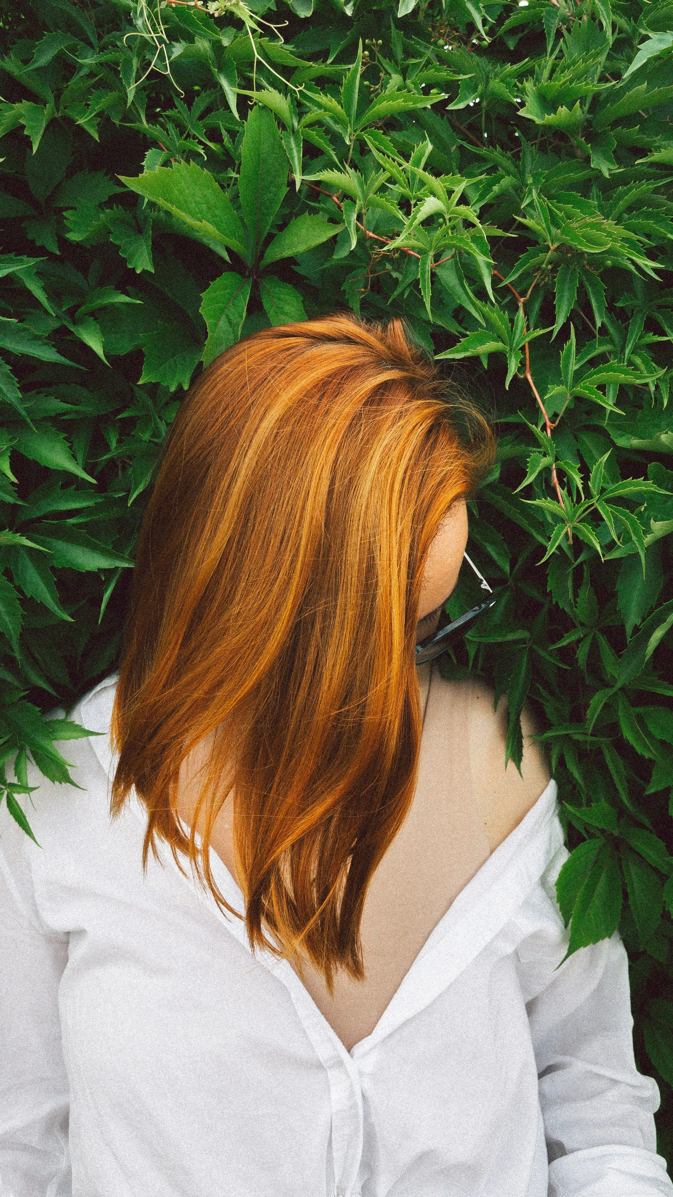 A woman with long, straight red hair wearing a white shirt, surrounded by lush green leaves.