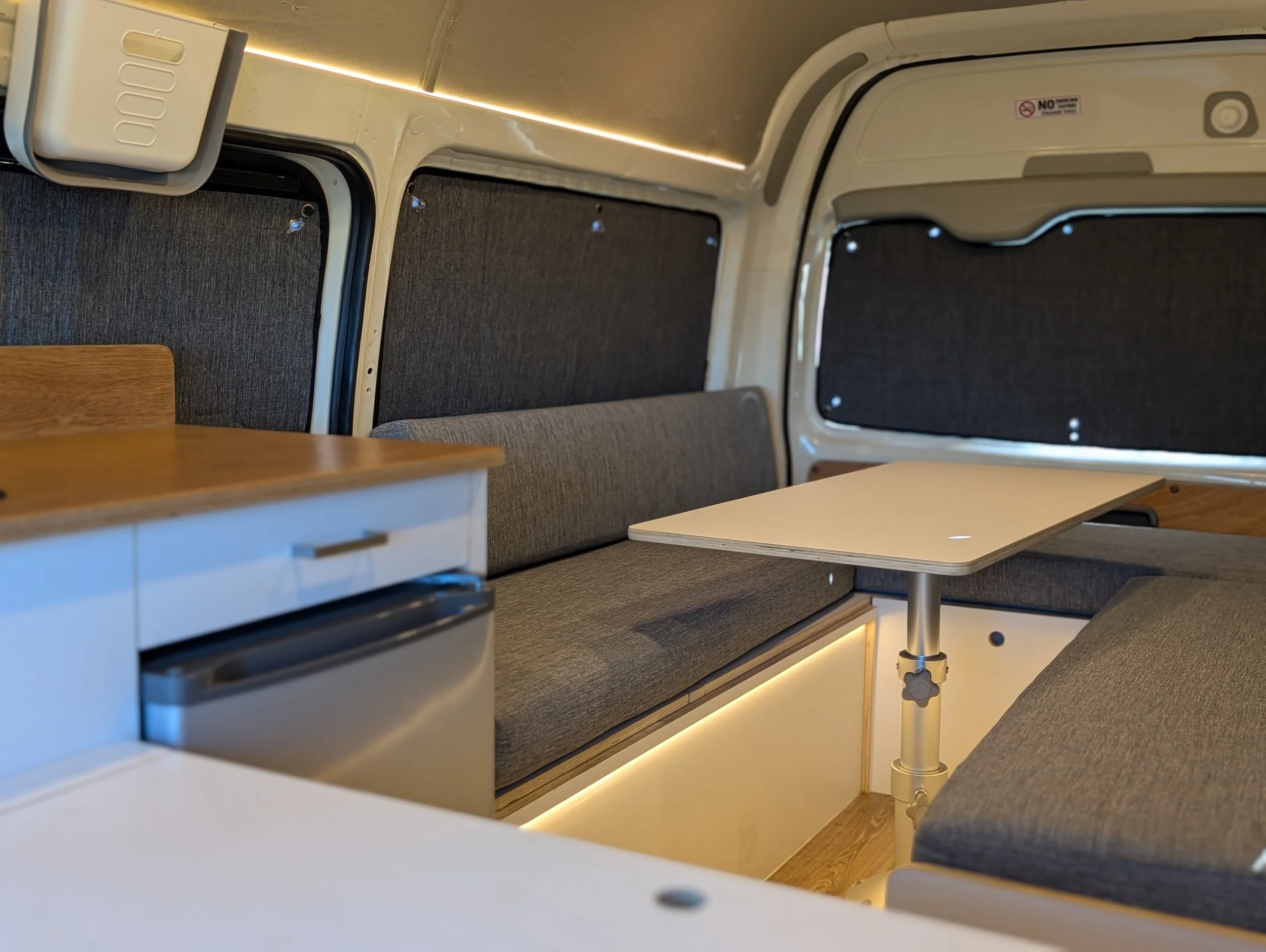 Interior of a modern camper van showing grey upholstered seats with tables, white walls, small windows with black curtains, and ambient lighting.
