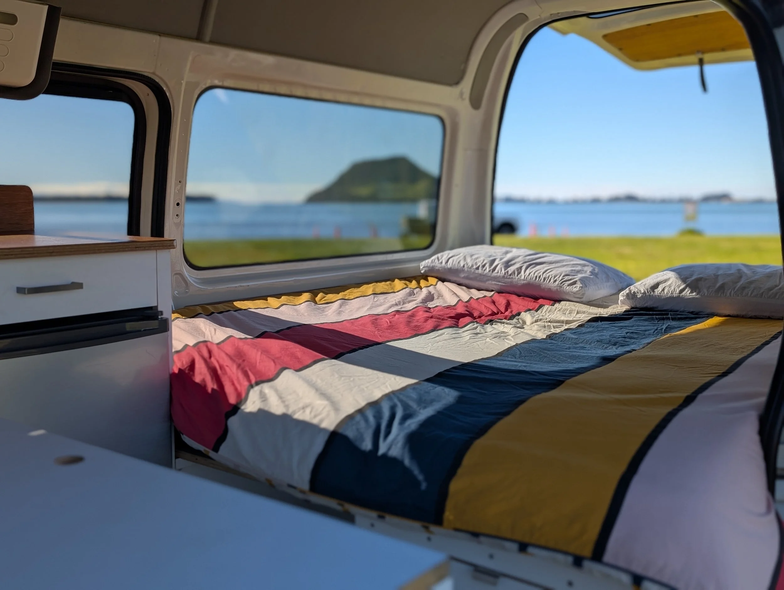 Interior of a camper van with a bed covered with a colorful striped blanket, pillows, and large windows showing a view of a body of water, mountains, and clear blue sky.
