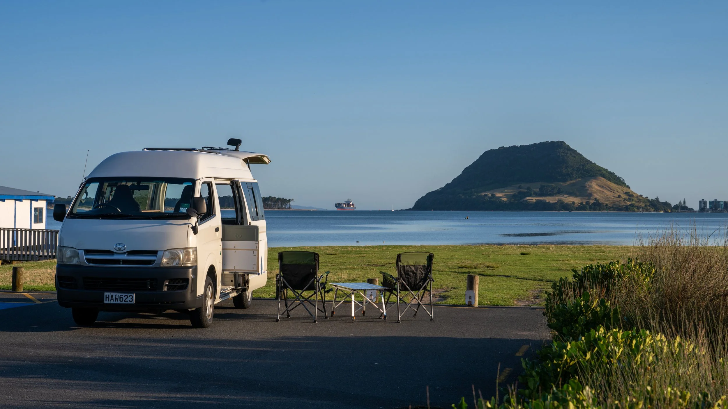 A white camper van parked near a lake with two camping chairs and a small table in front, with a scenic view of a hill surrounded by water and a ship in the distance under a clear blue sky.
