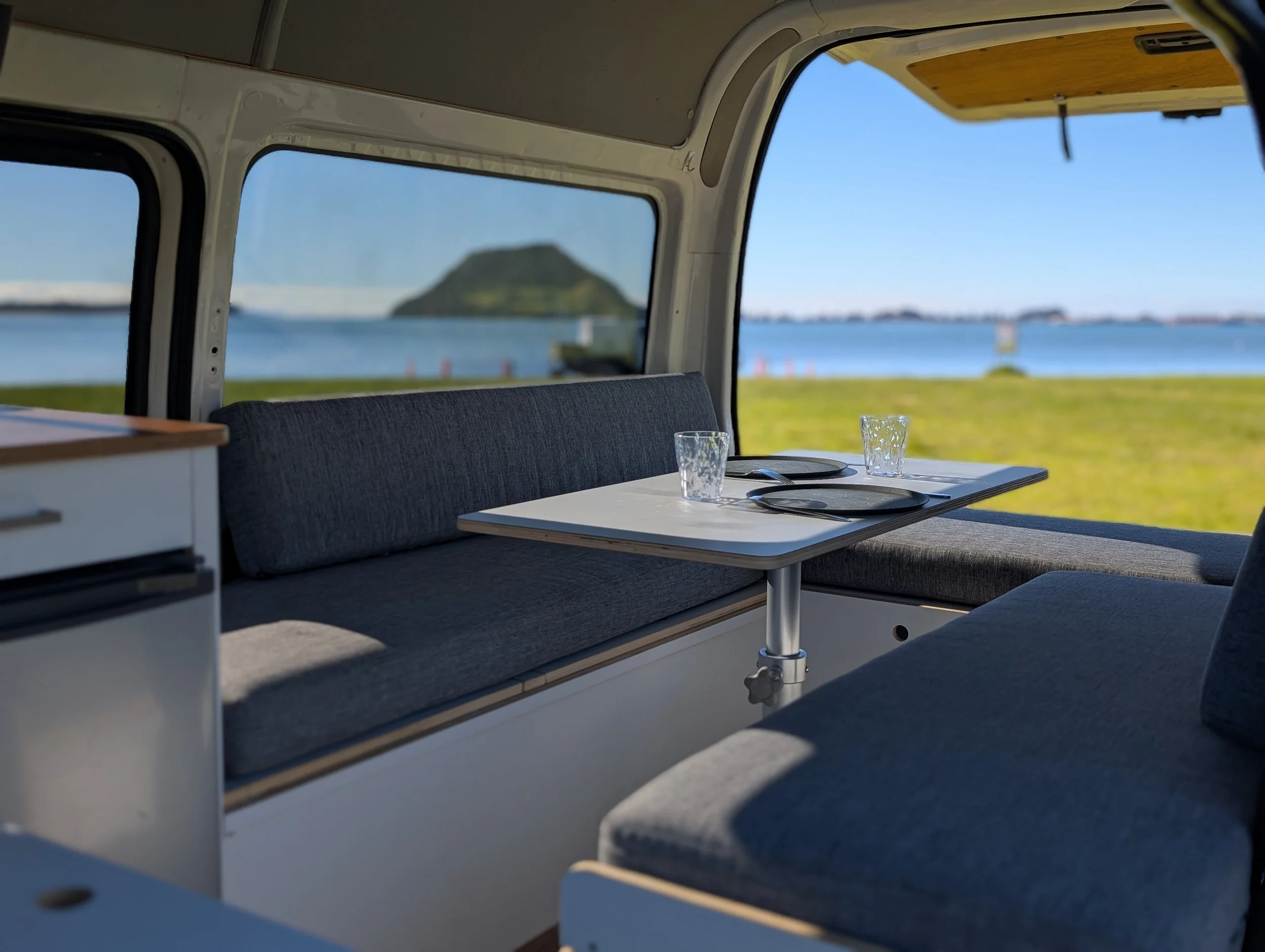 Interior of a motorhome or camper with a table set with glasses and plates, overlooking a scenic view of water and a distant island through large windows.
