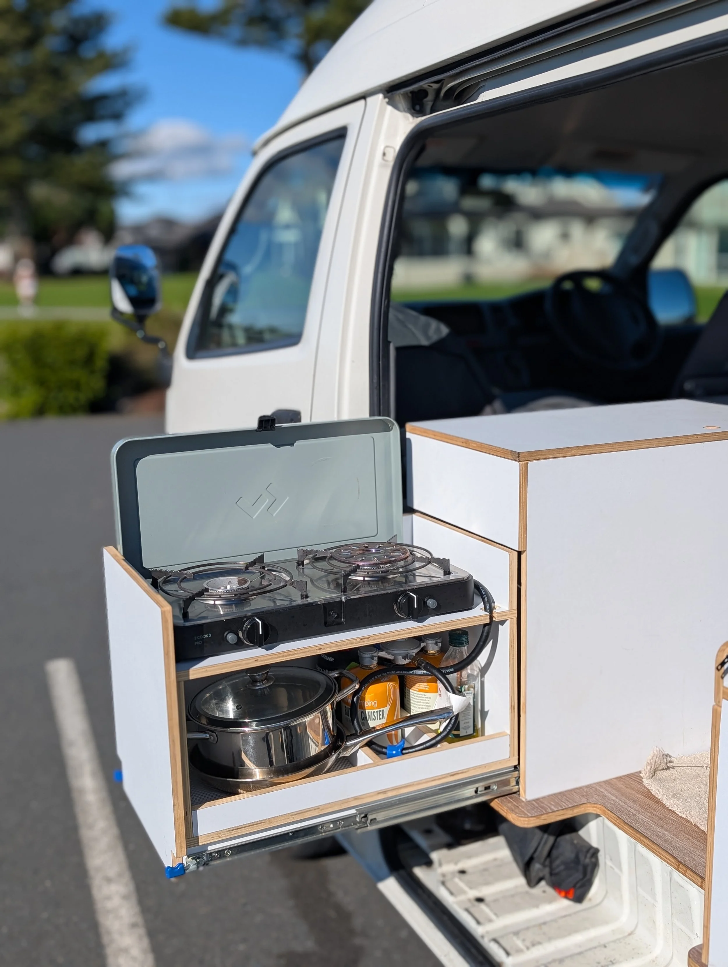 A small kitchen setup with a portable stove inside a wooden frame, installed in the back of a vehicle with an open door, showing kitchen supplies and bottles underneath, with a blurred outdoor scene in the background.