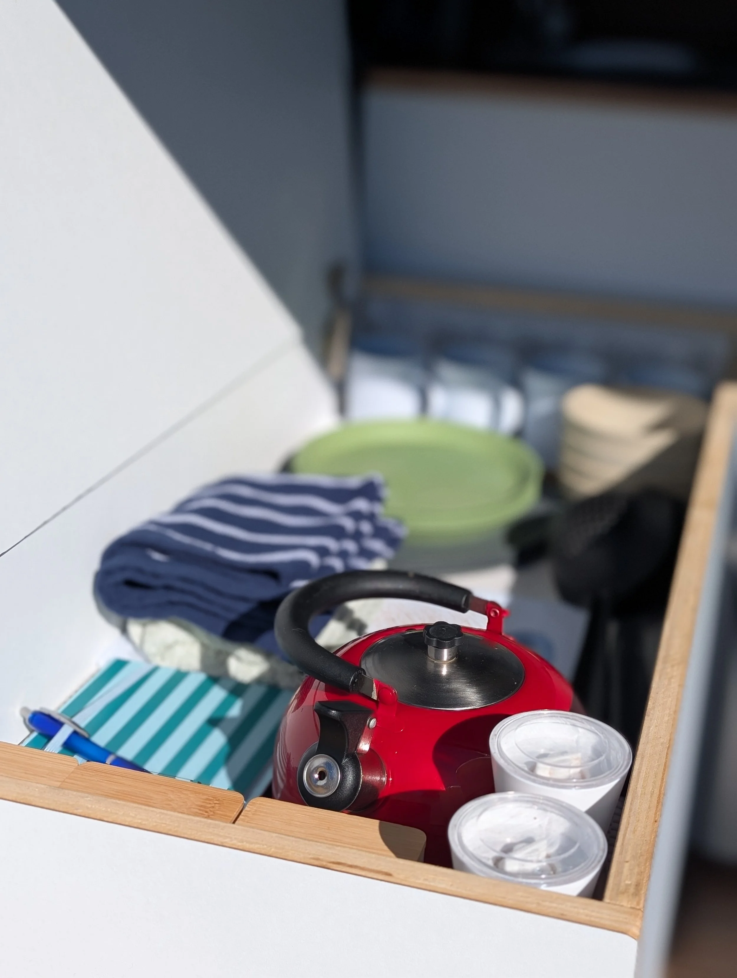 Wooden shelf with a red kettle, three containers, a striped cloth, and a blue and white pen. Blurry background showing a desk and chair.