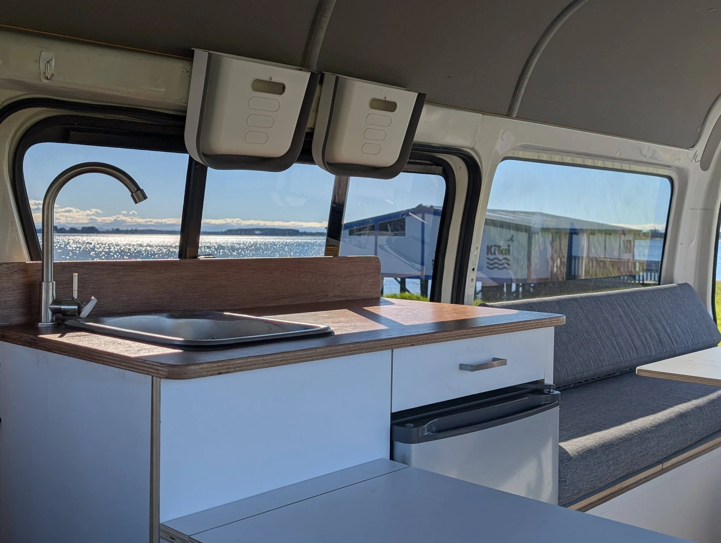 Interior of a RV with a small kitchen sink, faucet, and a wooden countertop, overlooking water and a distant shoreline through windows.