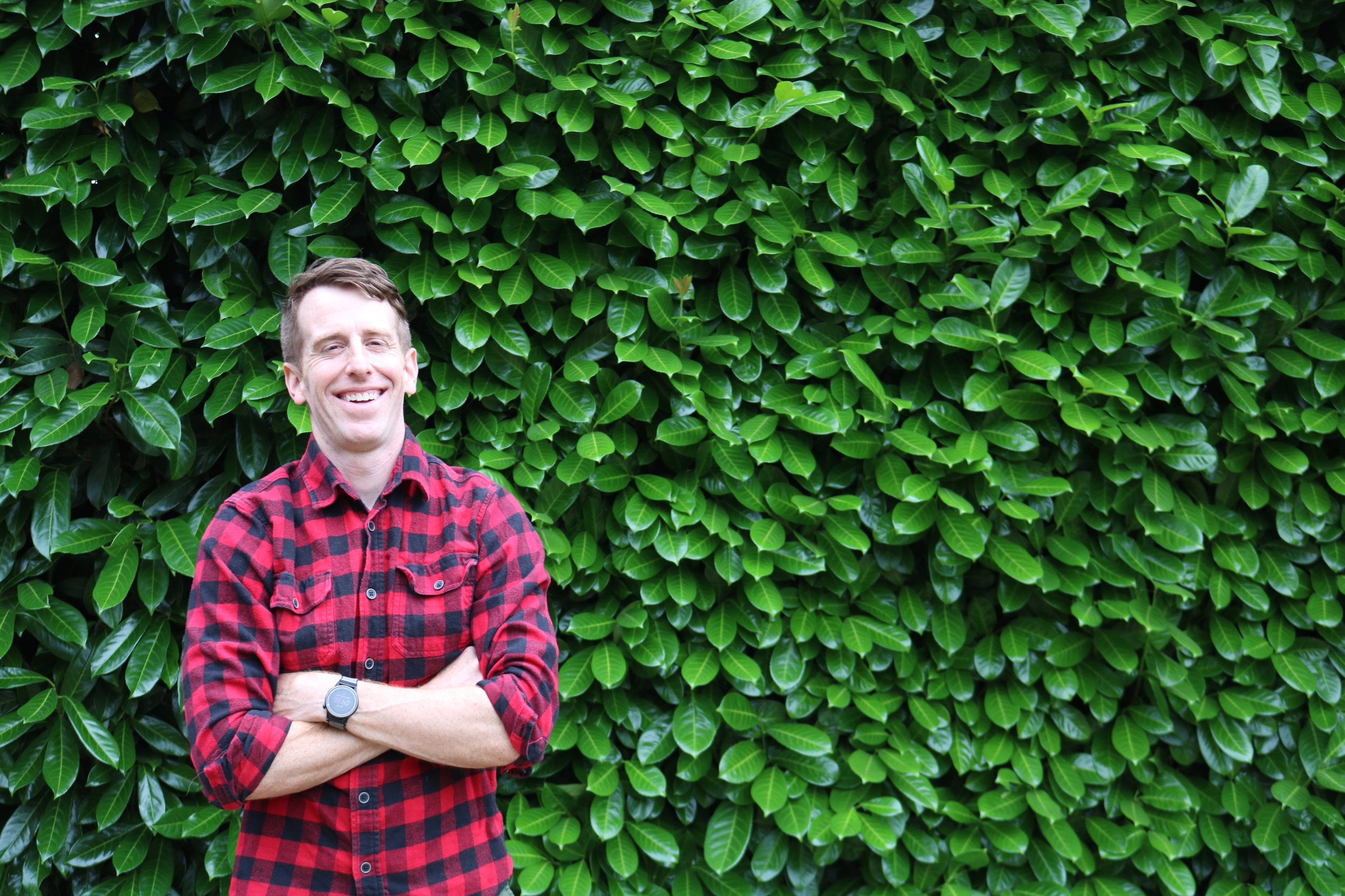 Dr. Dan Slowey, MD, wearing a red and black plaid shirt, smiling widely and standing in front of a lush green laurel hedge.