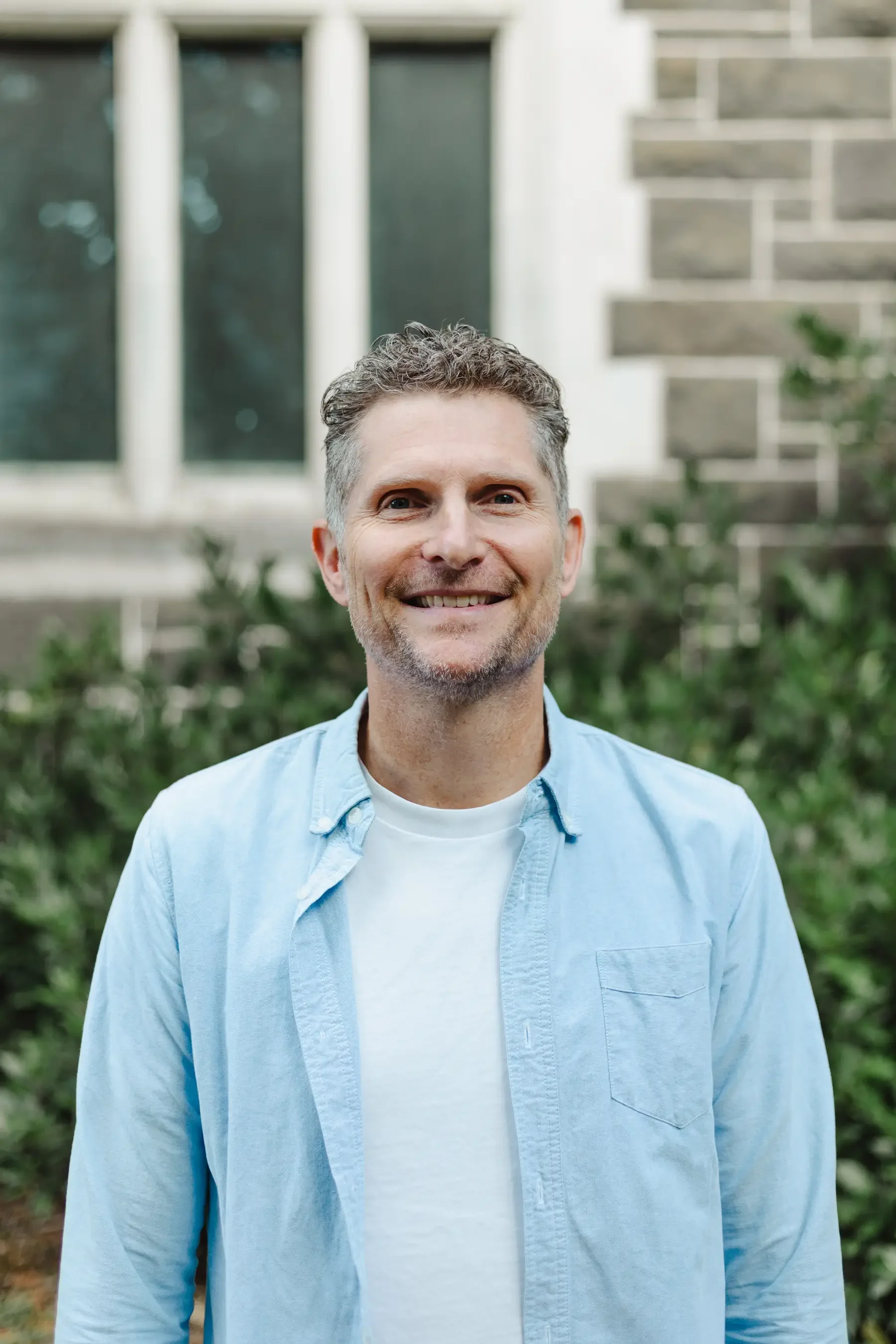 Rev Matt Bruns from Oasis Supervision standing in front of a stone building. He's wearing a white t-shirt with a light blue shirt over the top.