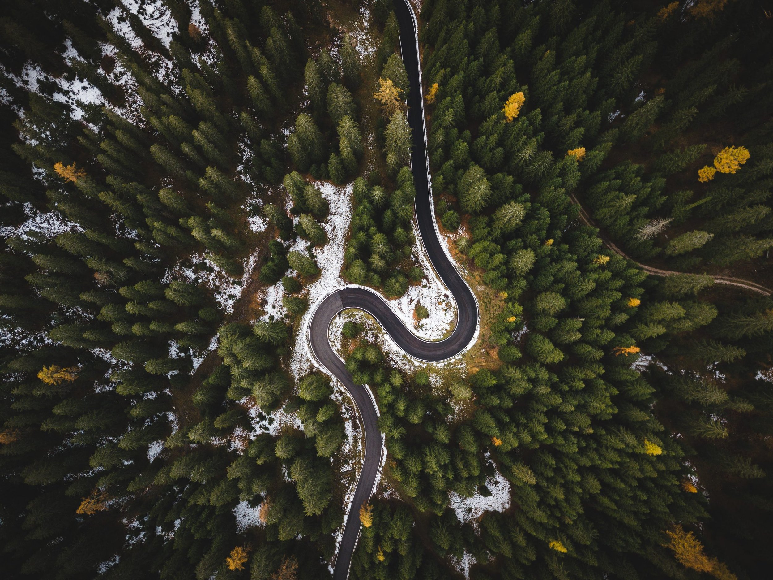 An aerial view of a winding road through a dense forest with patches of snow on the ground.