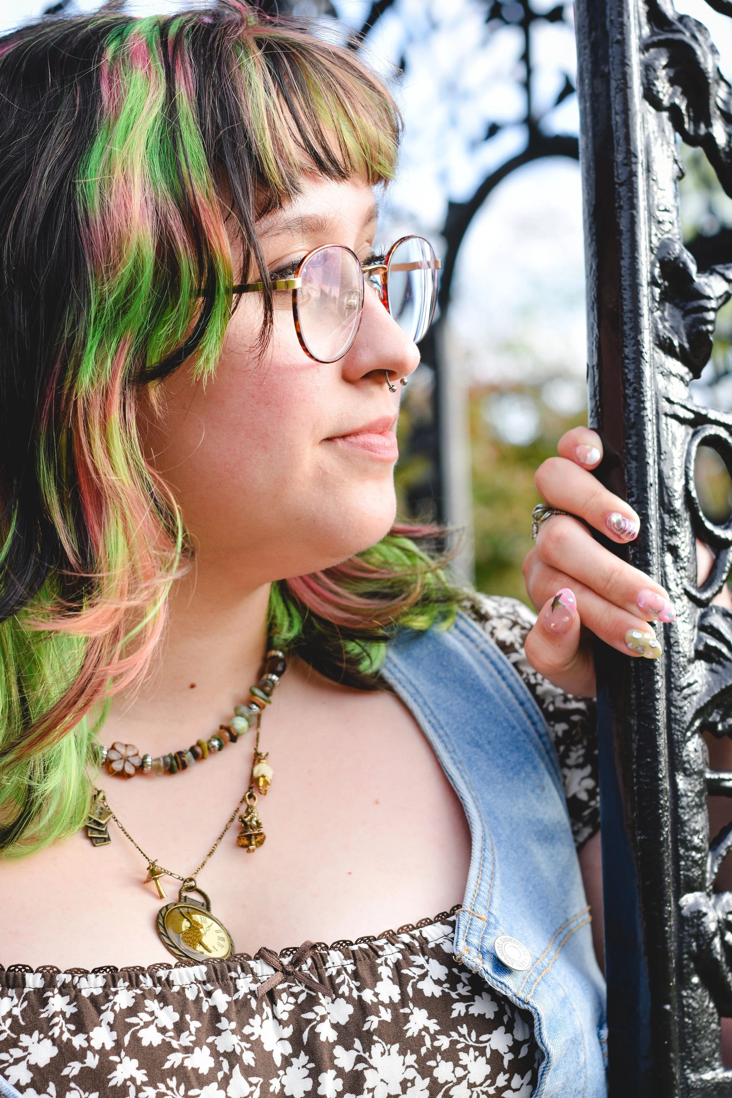 A young woman with colorful hair and glasses, wearing layered necklaces and a floral-patterned dress with a denim vest, holding onto a black iron fence outdoors.