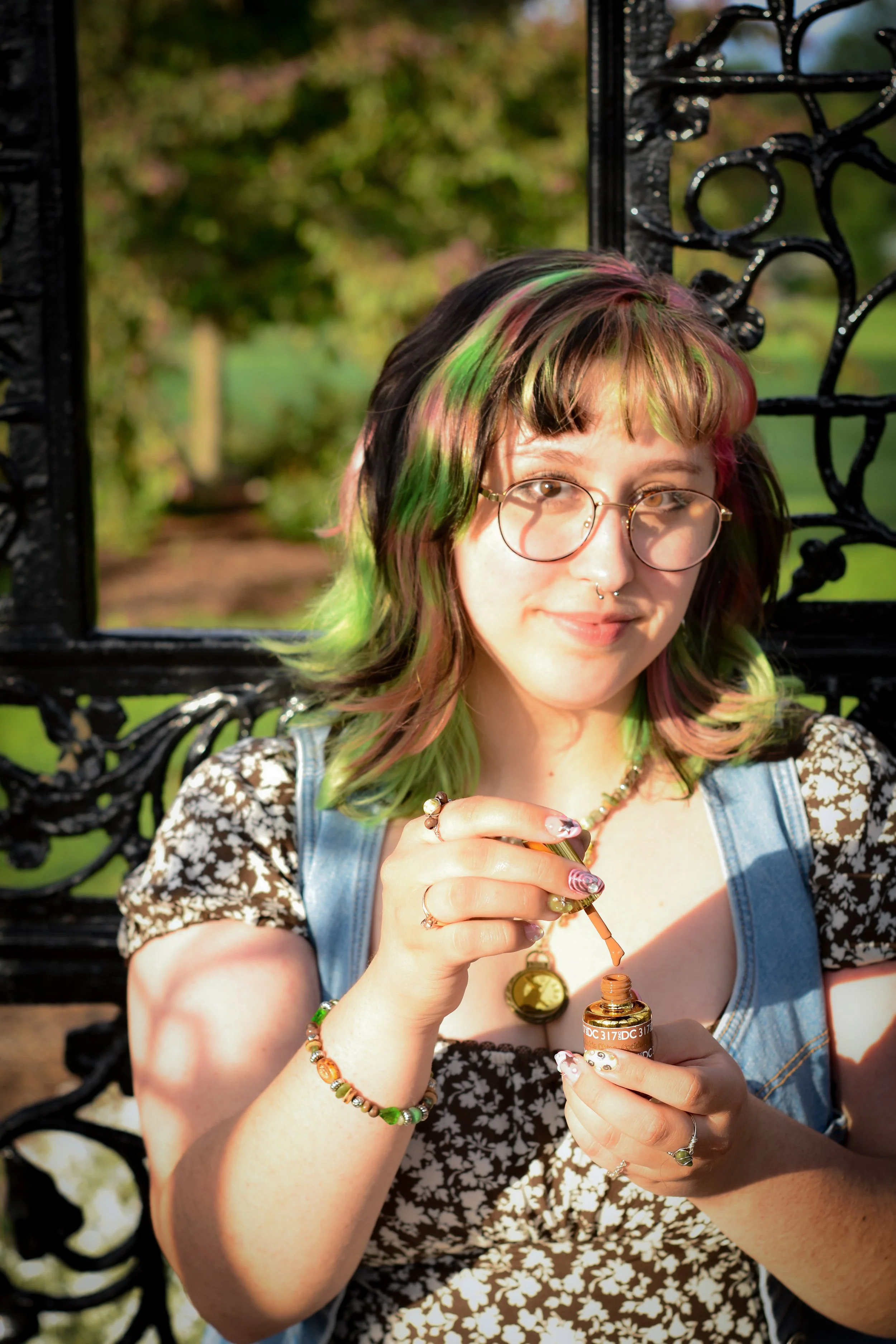 A young woman with multi-colored hair, wearing glasses, sitting on a black ornate park bench in a park, holding and opening a small amber vial of ointment or medication.