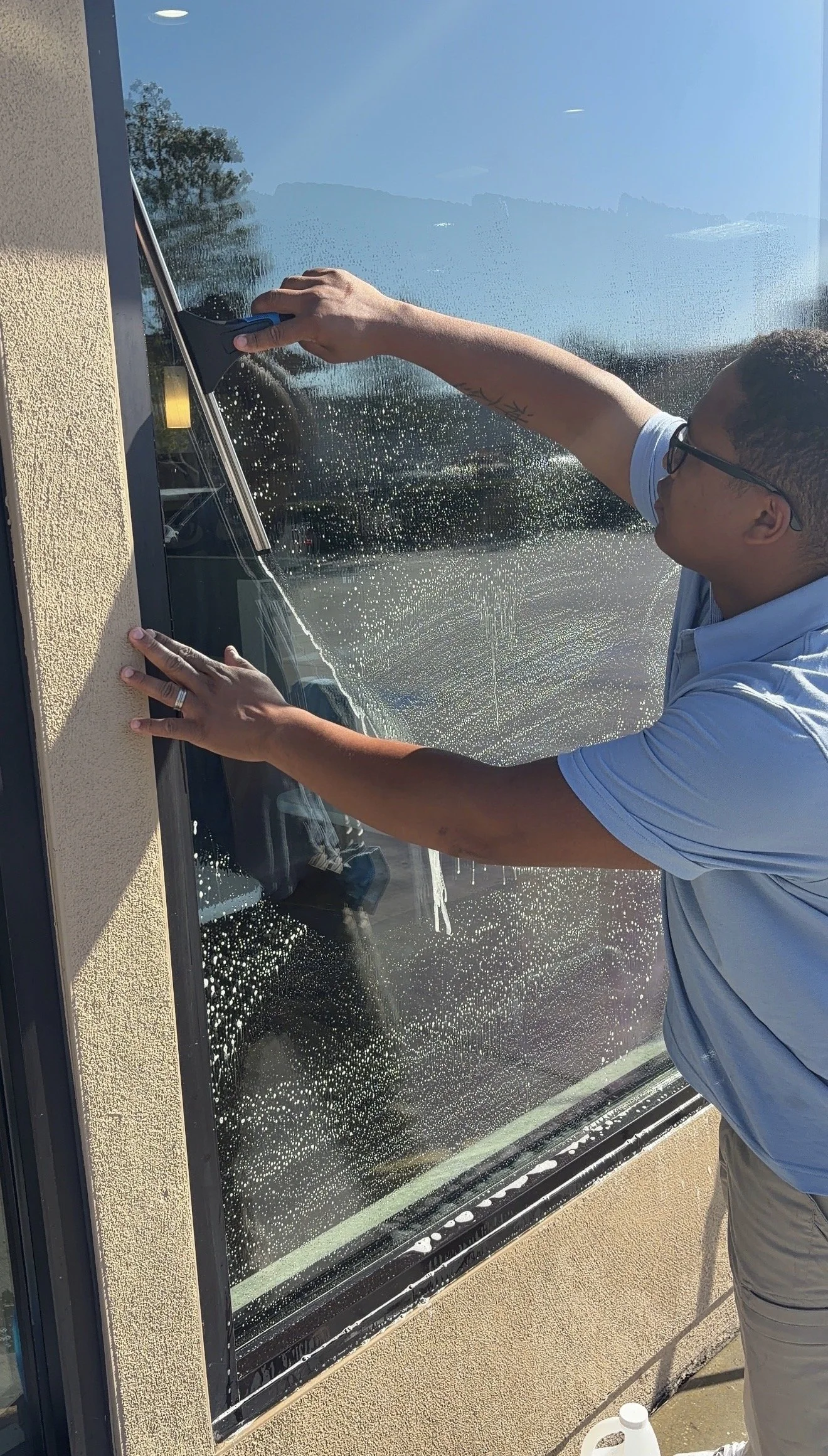 Person cleaning a large glass window with a squeegee on a sunny day, with the reflection of trees and sky visible on the window.