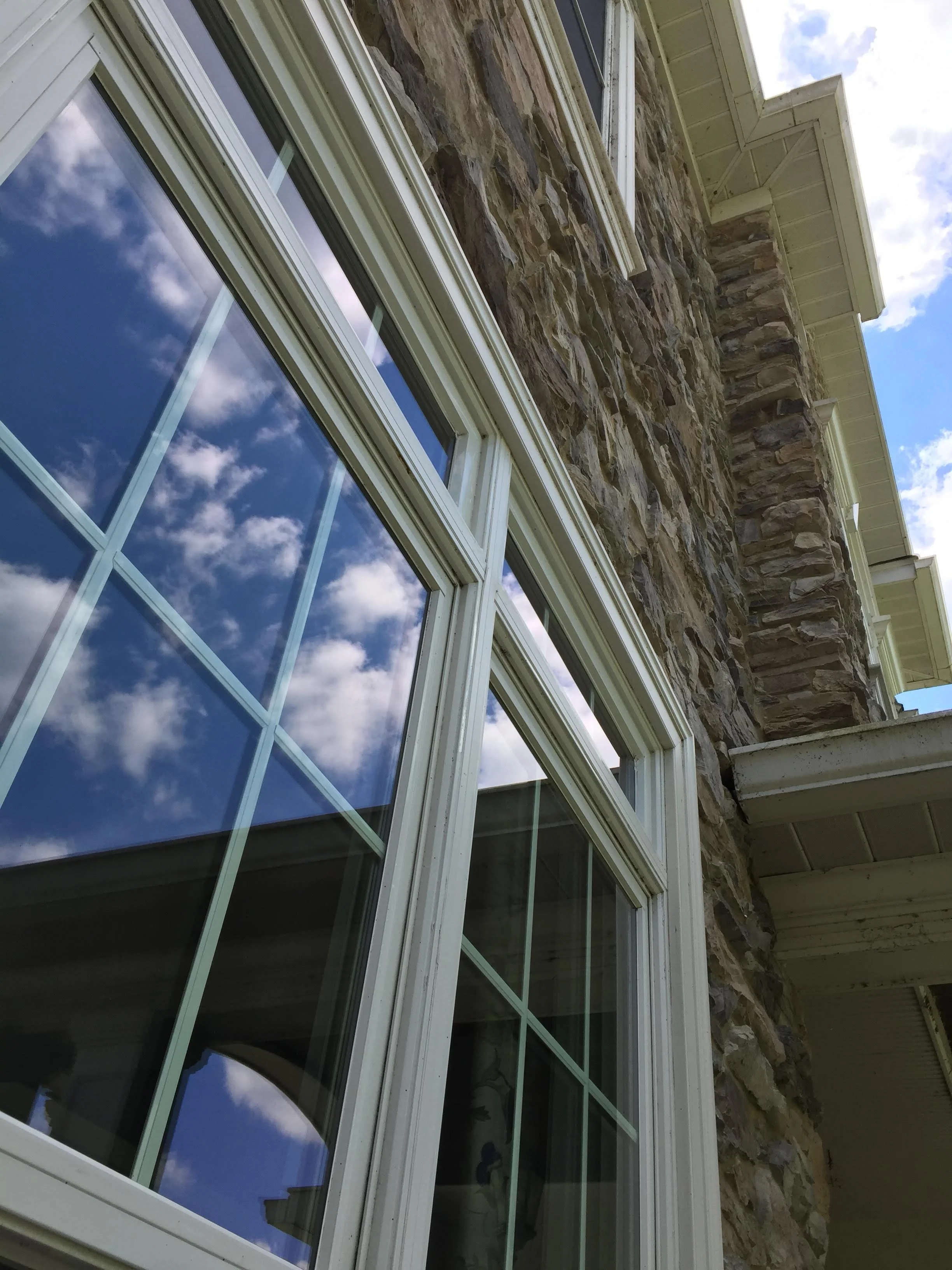 View of house exterior showing large multi-pane windows reflecting the sky and clouds, stone wall facade, and part of the roof's soffit.