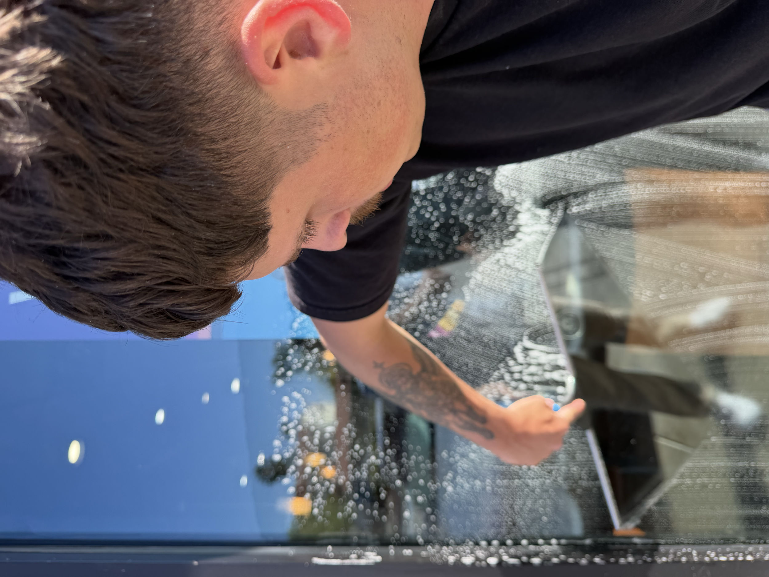 A person with dark hair and a tattoo on their arm, leaning over a glass surface in an outdoor setting, with a blue sky visible in the background.