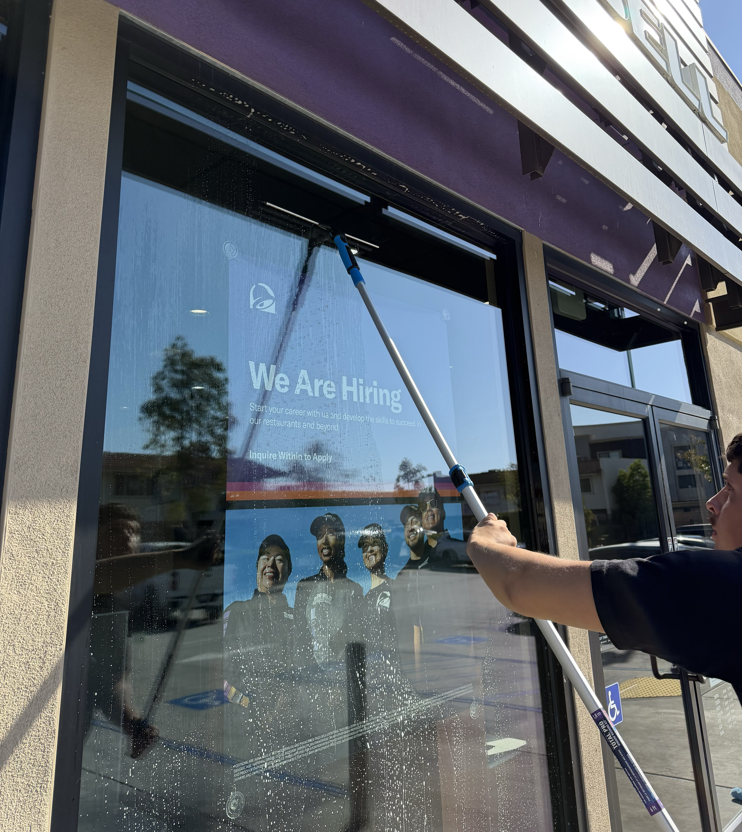 A person using a squeegee to clean a glass door with a 'We Are Hiring' sign on it, outside of a building with a beige wall and windows.