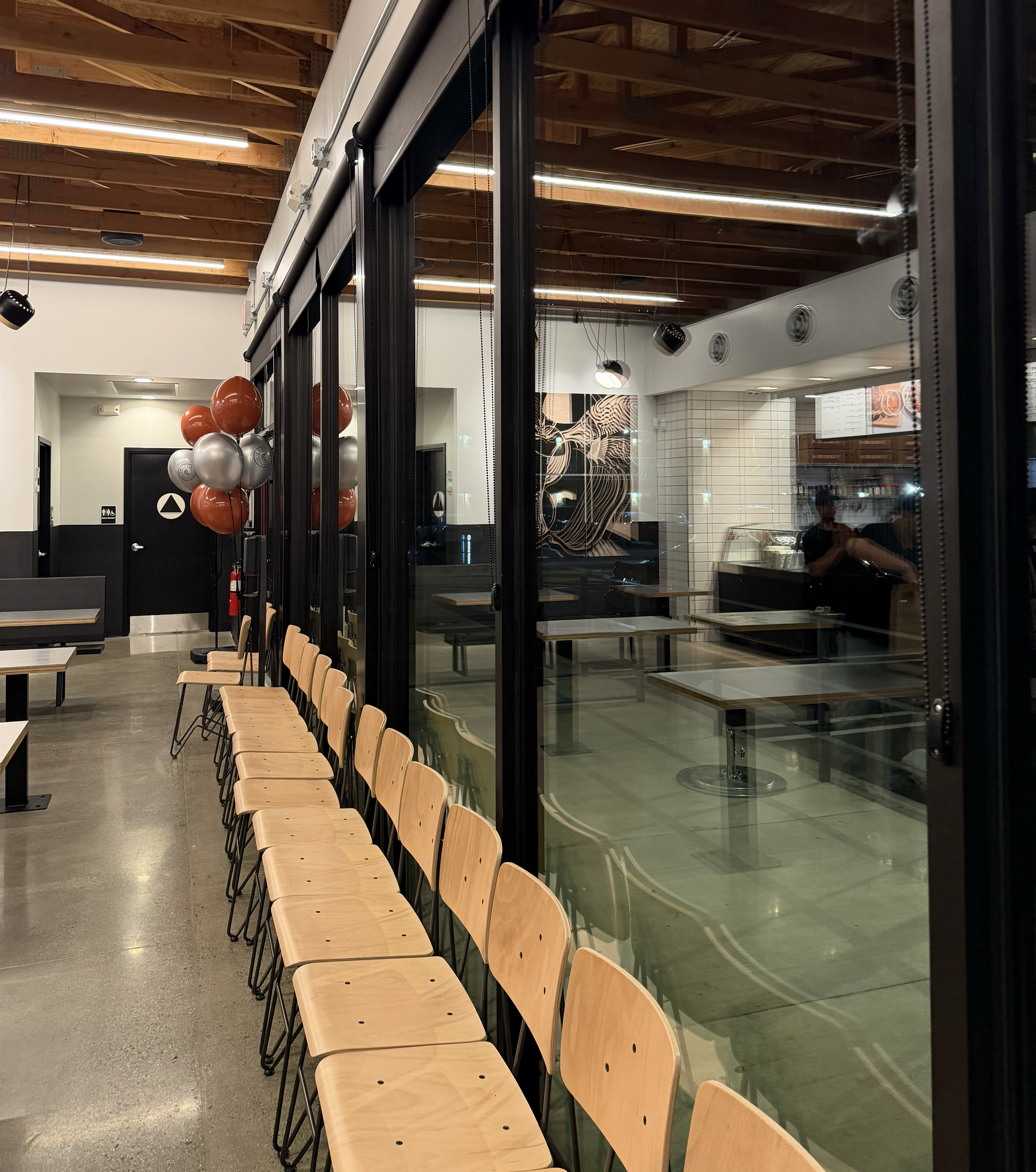 Interior of a modern restaurant or cafe with a row of wooden chairs lined up along a glass wall, balloons in red, silver, and white colors, and a partially visible menu board inside.