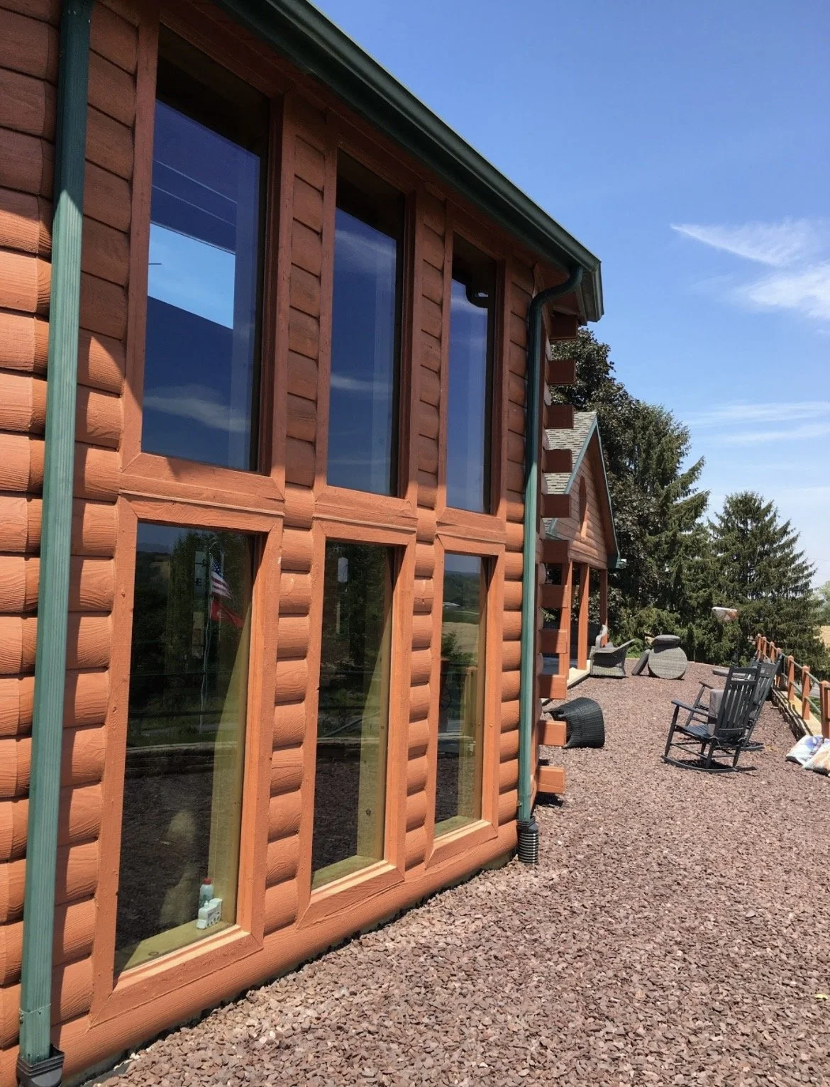 Exterior view of a log cabin with large windows, outdoor seating, and a gravel patio area, surrounded by trees on a bright, sunny day.