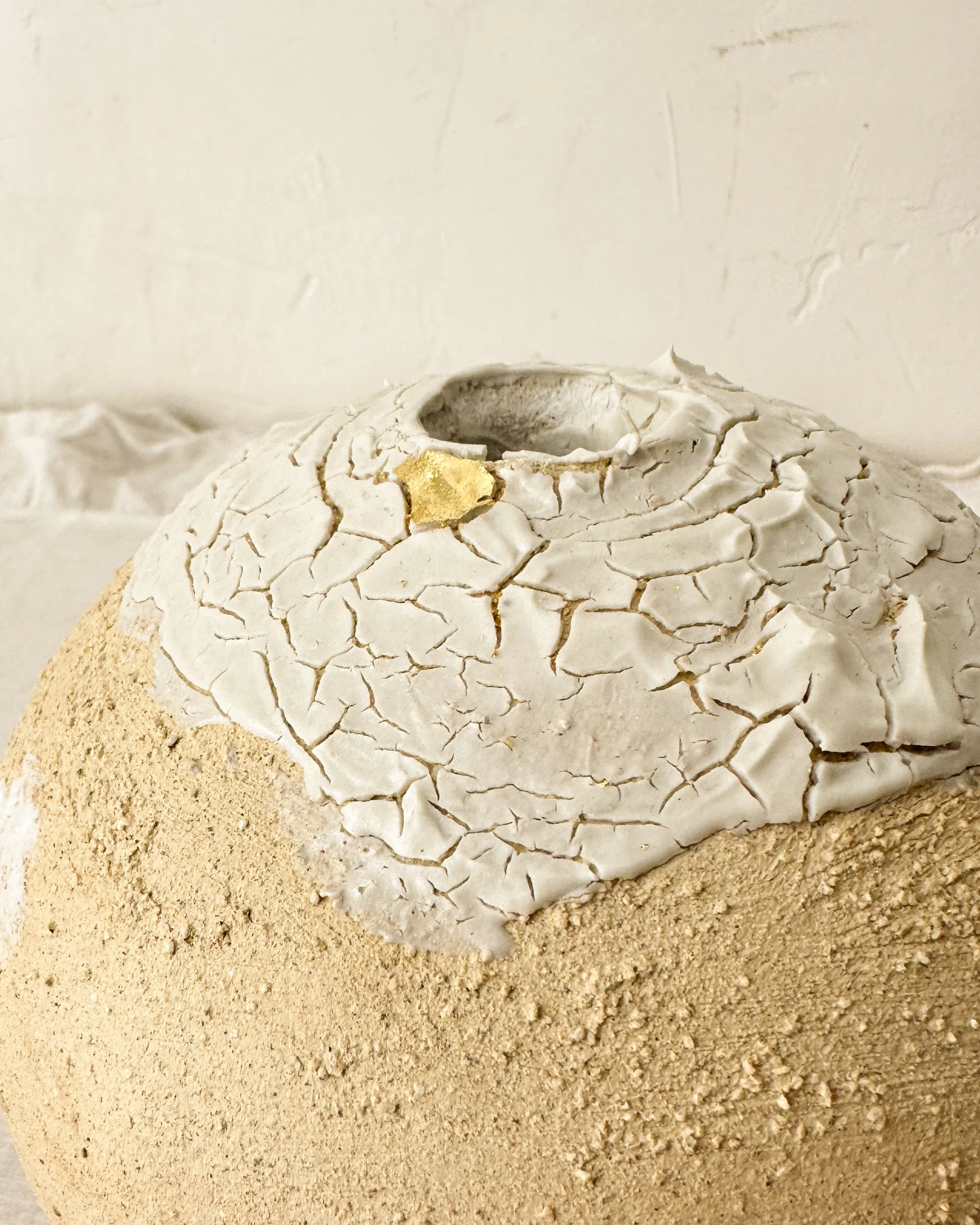 Une sculpture en forme de tête, faite de matériaux craquelés blanc et beige, avec une partie ornementale en or. La sculpture repose sur du sable.
