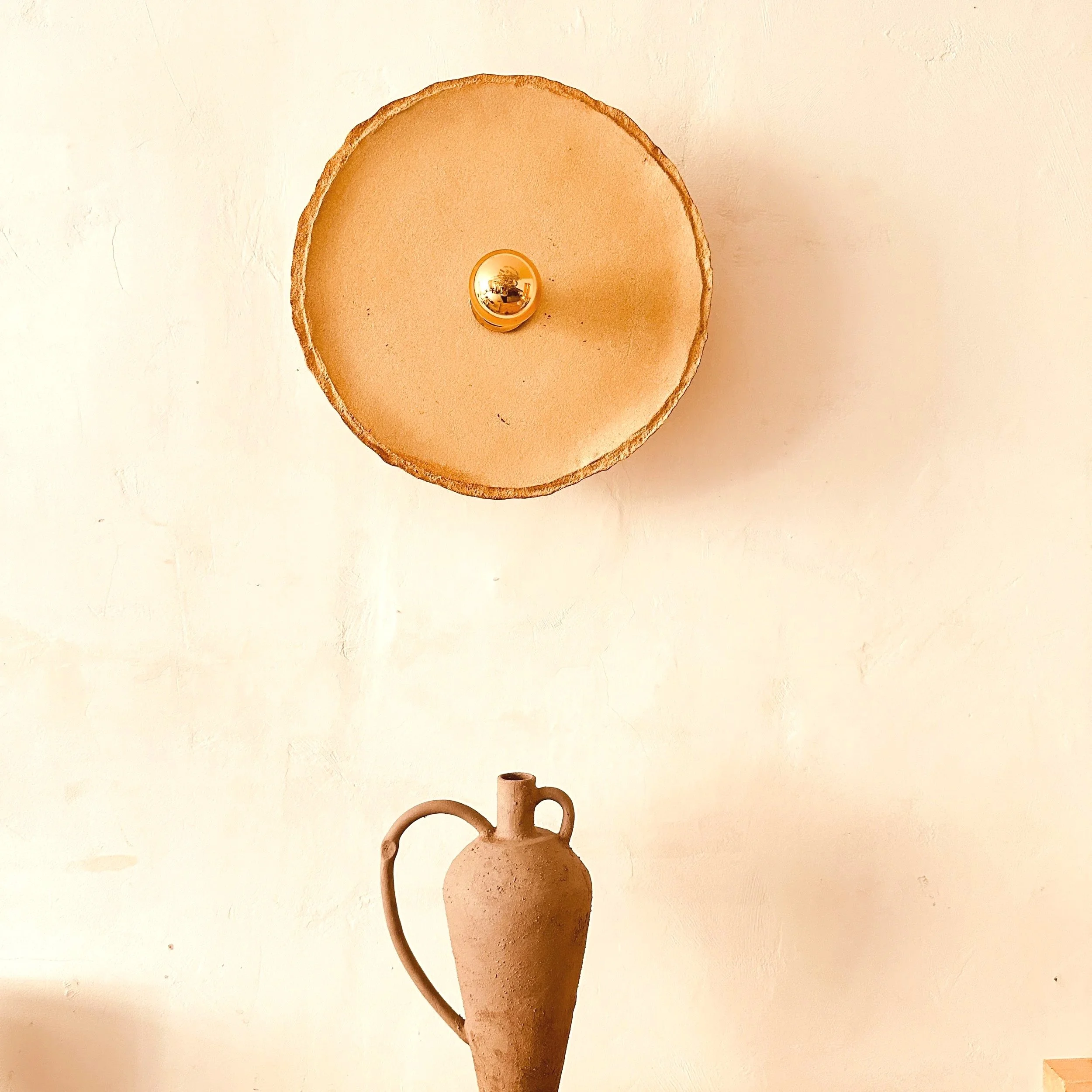 Une sculpture en terre cuite représentant un vase ou un objet à forme organique avec une tige ou une prise, placé devant un mur blanc.