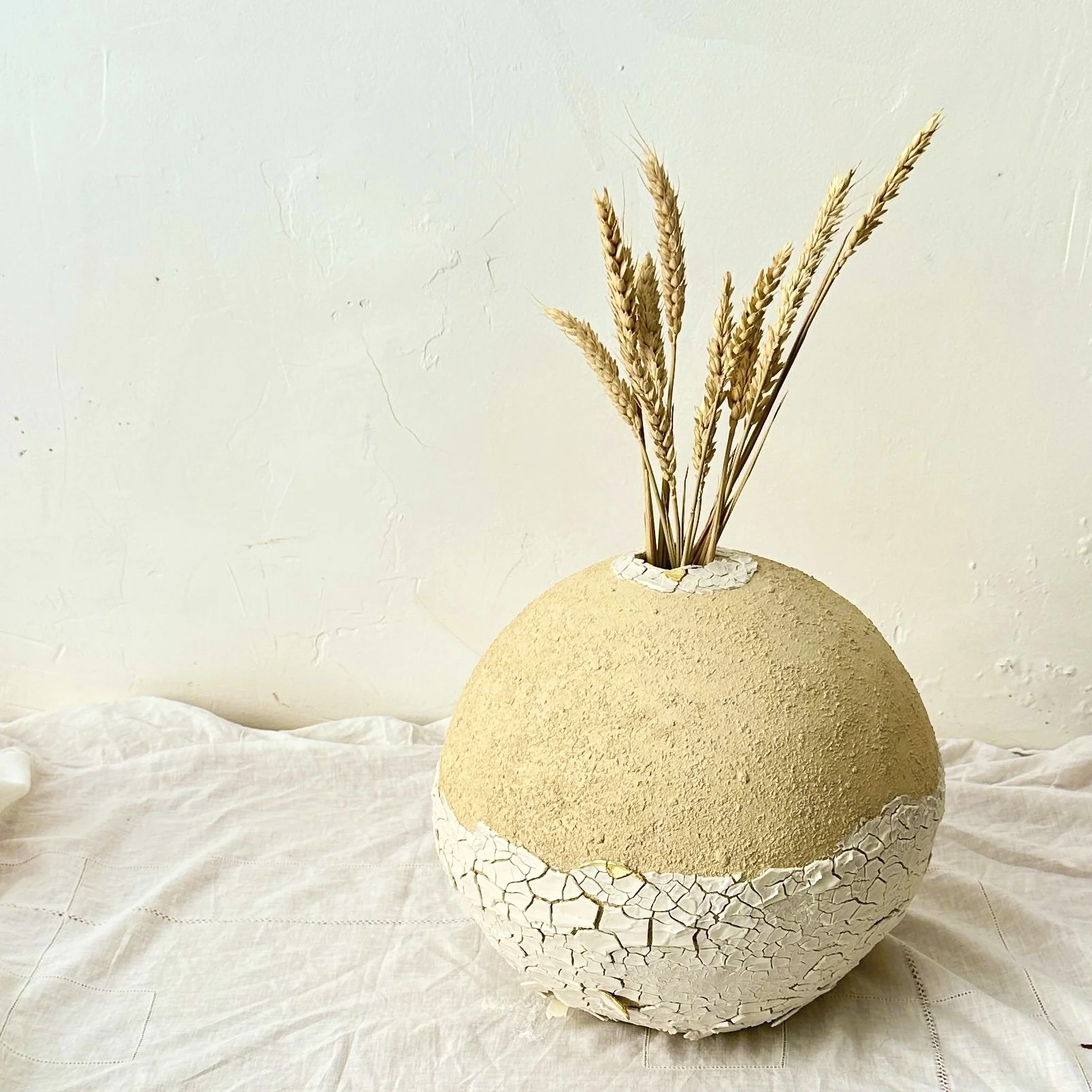 Vase rond blanc avec surface craquelée, contenant des tiges de blé séchées, posé sur une table en tissu blanc, fond blanc
