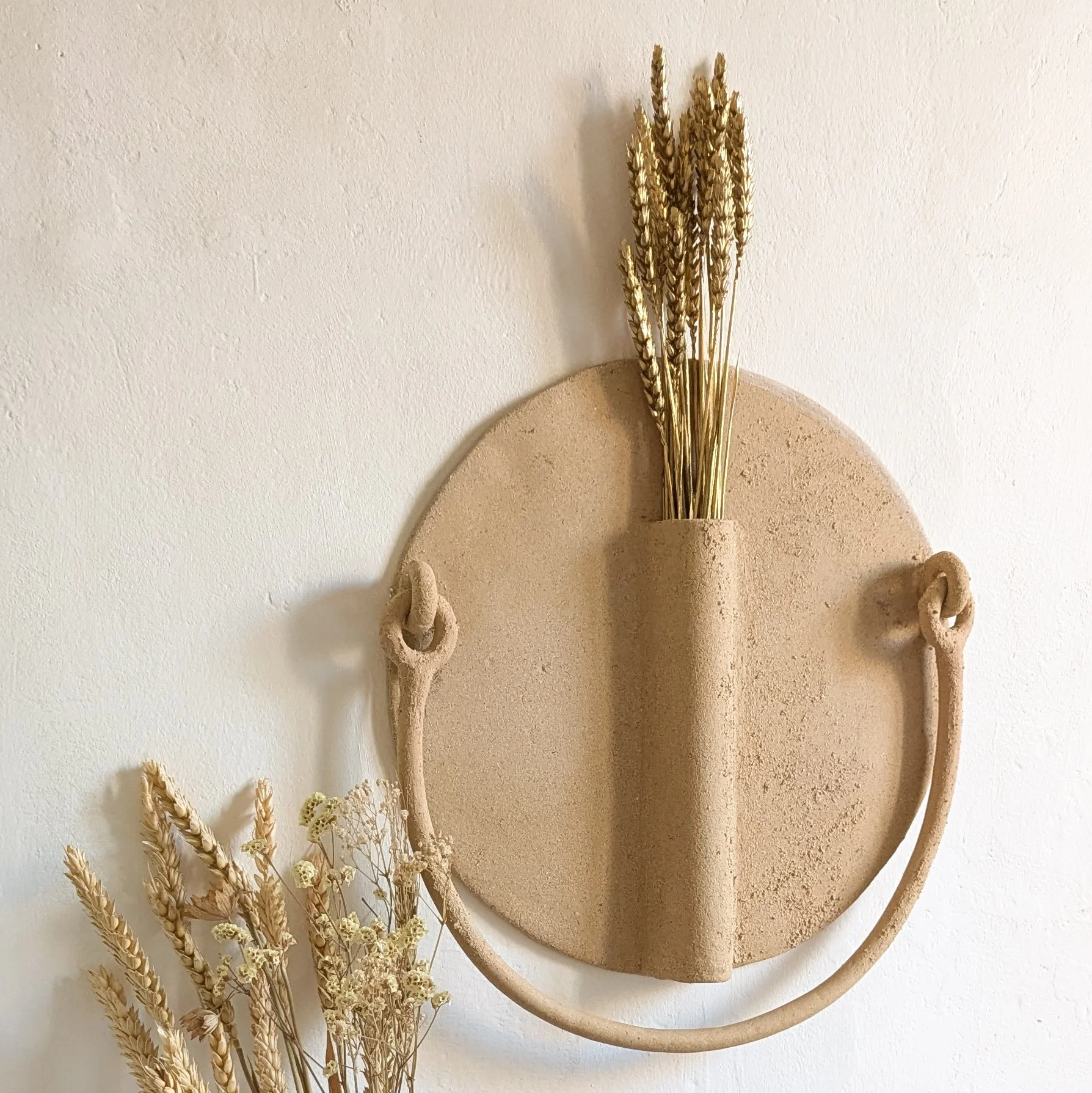 Une décoration murale en formes ovales en terre cuite avec des branches de céréales séchées suspendues, décorée avec des fleurs séchées en bas à gauche.