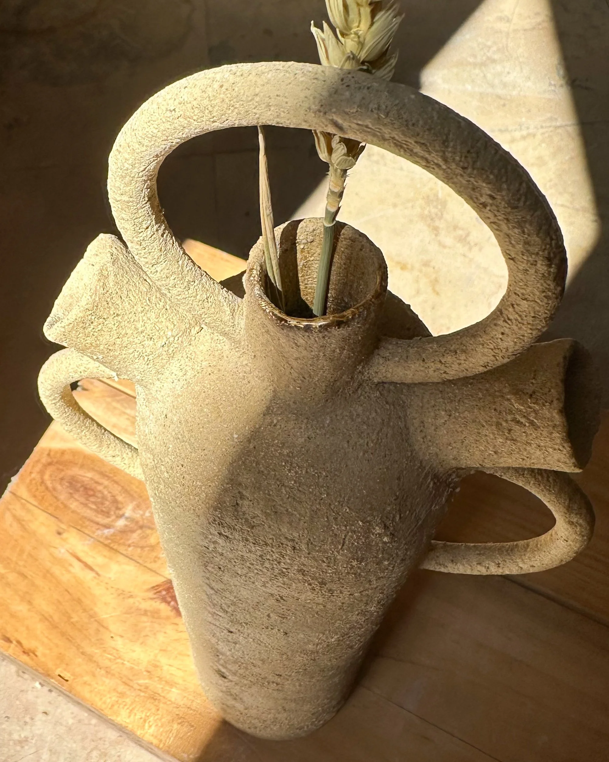 Une sculpture en forme de spirale en pierre avec une ouverture centrale, contenant des brins d'herbe. La sculpture est placée sur une table en bois, avec un fond en pierre et un sol en carrelage.