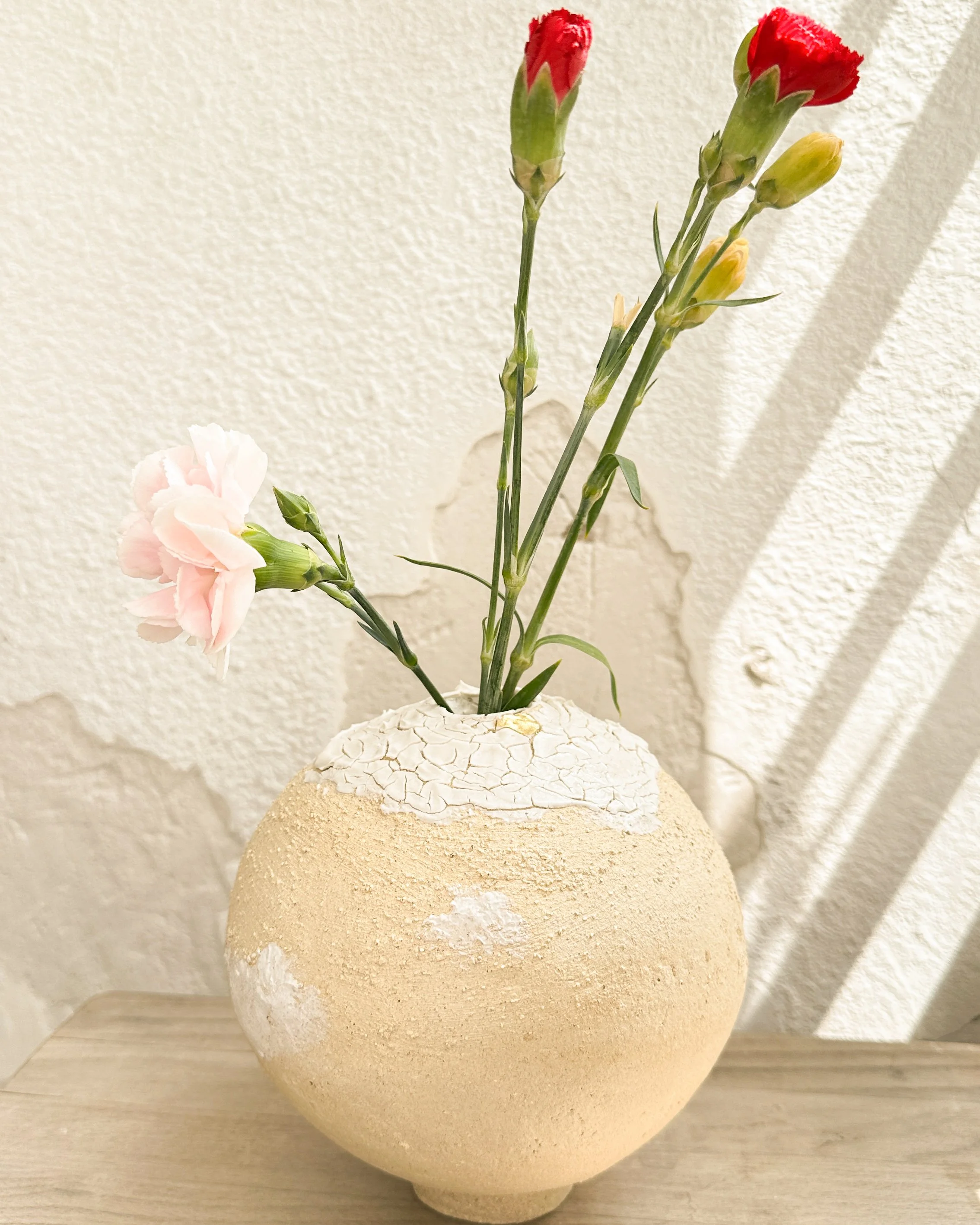 Un vase blanc et beige avec une surface rugueuse, contenant des fleurs aux pétales roses et à d'autres fleurs à tiges vertes avec des bourgeons. Le vase est placé sur une surface en bois clair, en face d'un mur blanc texturé.