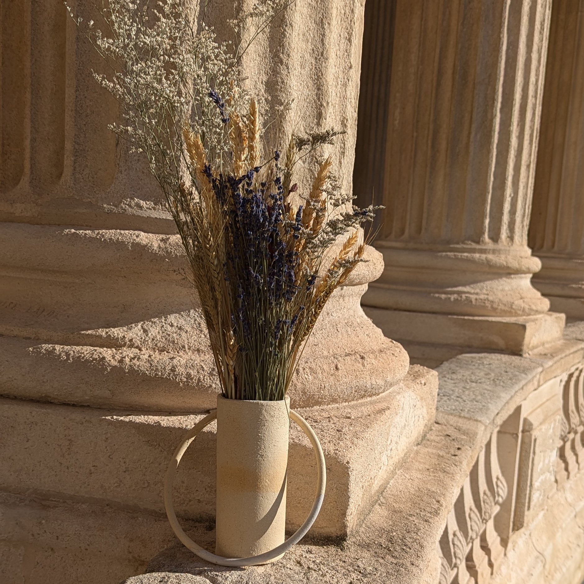 Un vase blanc contenant des bouquets de fleurs séchées, placé contre un mur en pierre beige avec des moulures.