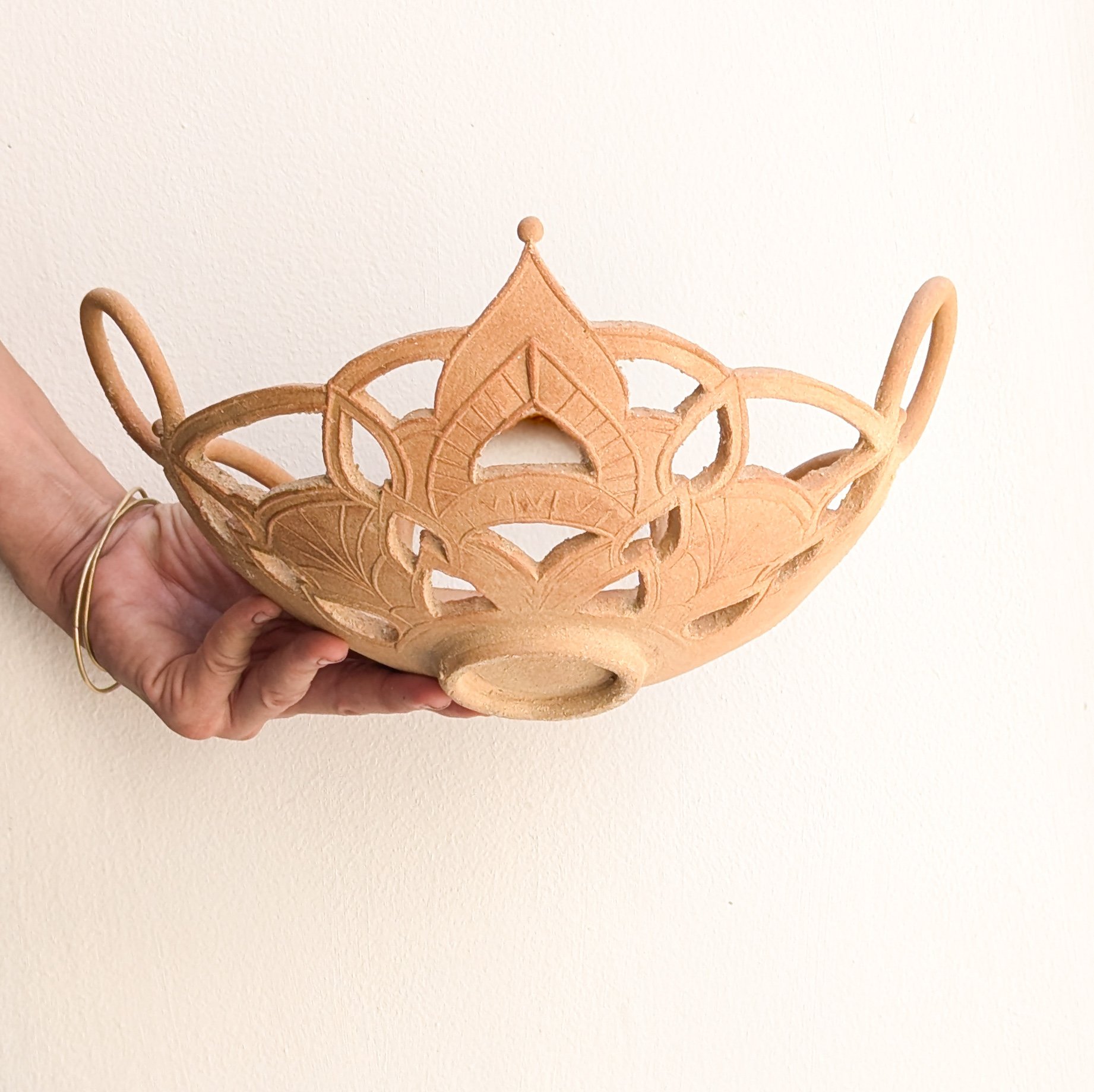 Un panier en argile ou en bois sculpté, avec un motif de couronnes, tenu par une main. Le fond est clair et un mur blanc.