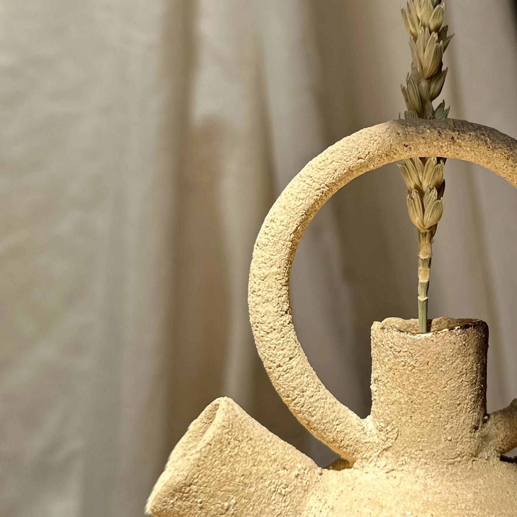 Une sculpture abstraite en forme de spirale en matériau beige, avec une tige végétale à l'intérieur, sur fond neutre.