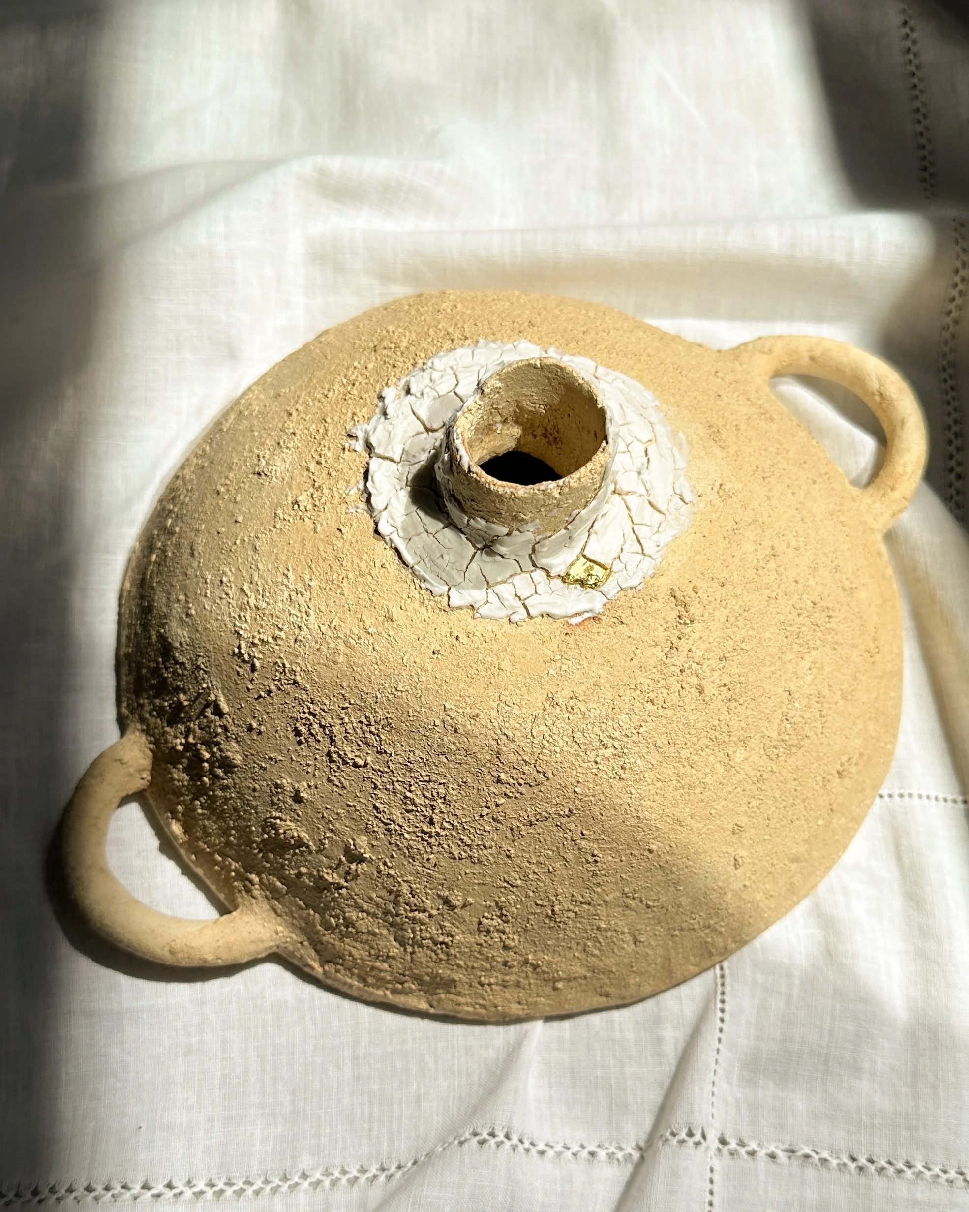 Une théière en terre cuite avec un couvercle en céramique blanche, placée sur une nappe en tissu blanc avec des broderies.