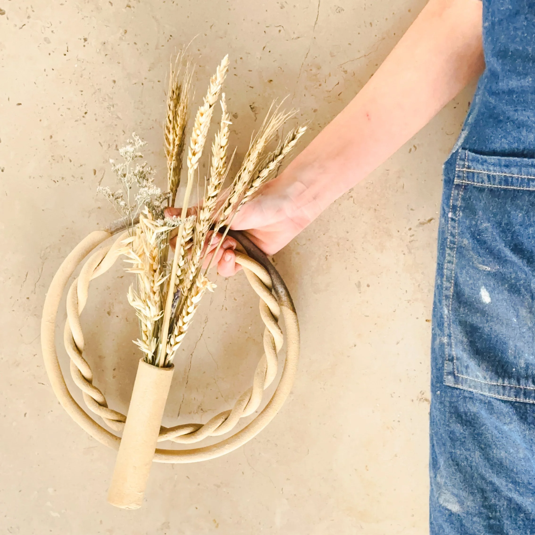Une main tenant un bouquet de blé séché, dans un porte-fleuvre, posé sur une surface en pierre ou en béton.