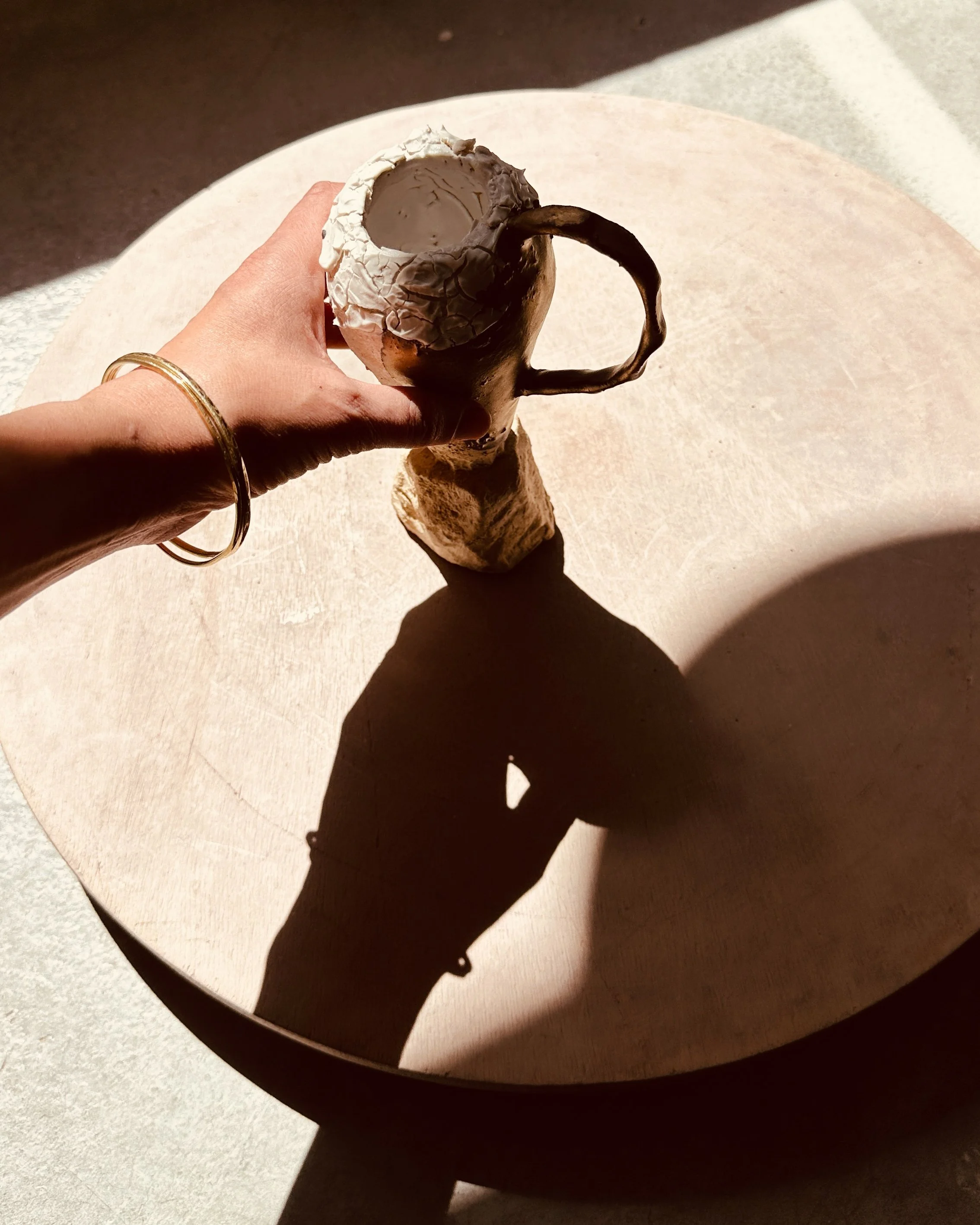 Une main tenant une tasse en forme de tête de cerf, garnie de crème et de chocolat, positionnée sur une surface beige avec des ombres contrastées.