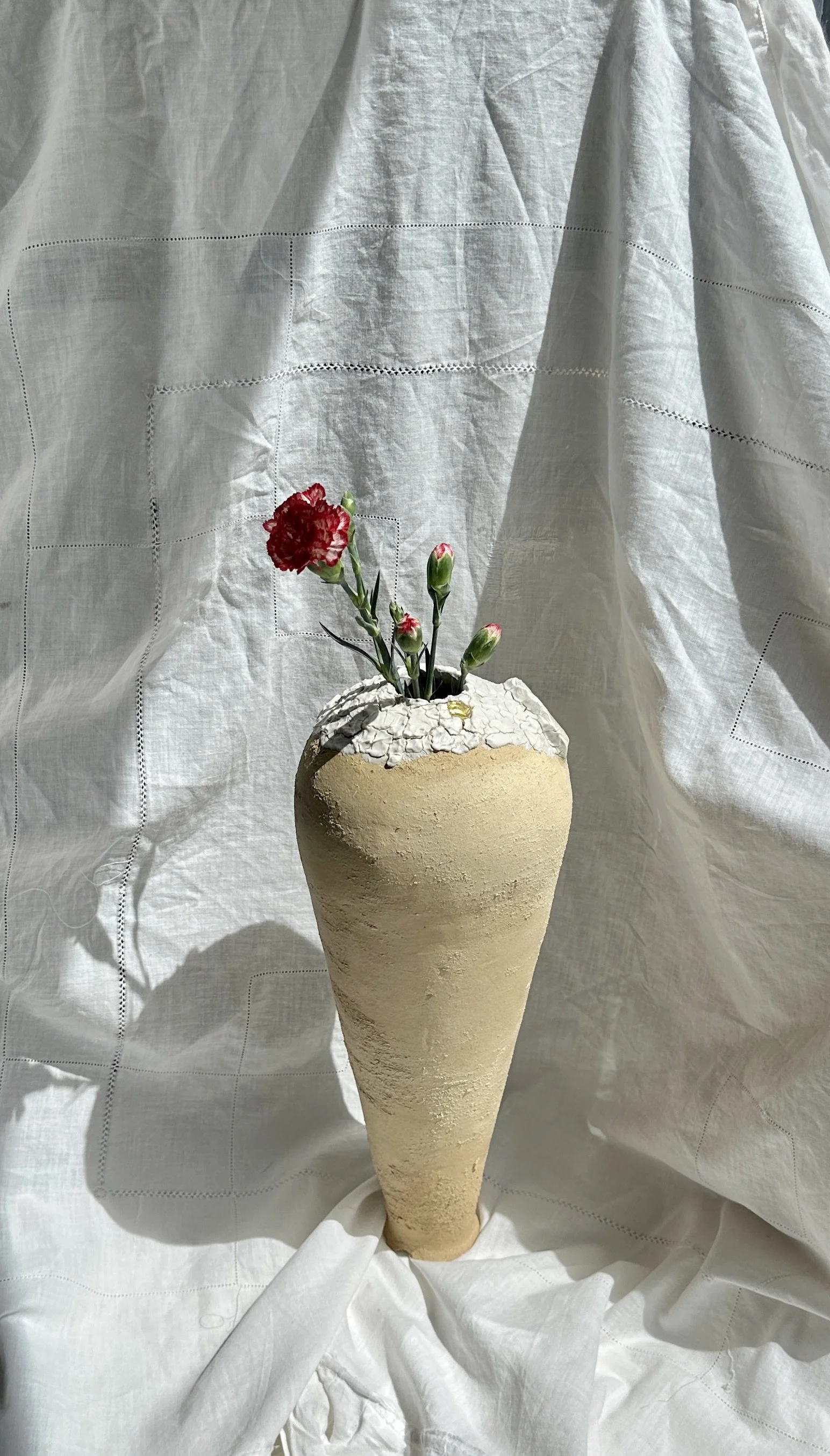Vase en forme de cône, avec des fleurs rouges et des bourgeons, posé devant un tissu blanc avec des détails de couture.