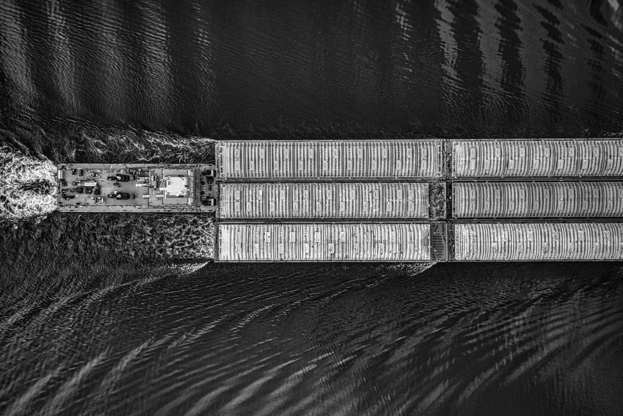 Aerial black and white photo of a cargo ship moving through water, with multiple containers on deck and wake at the stern.