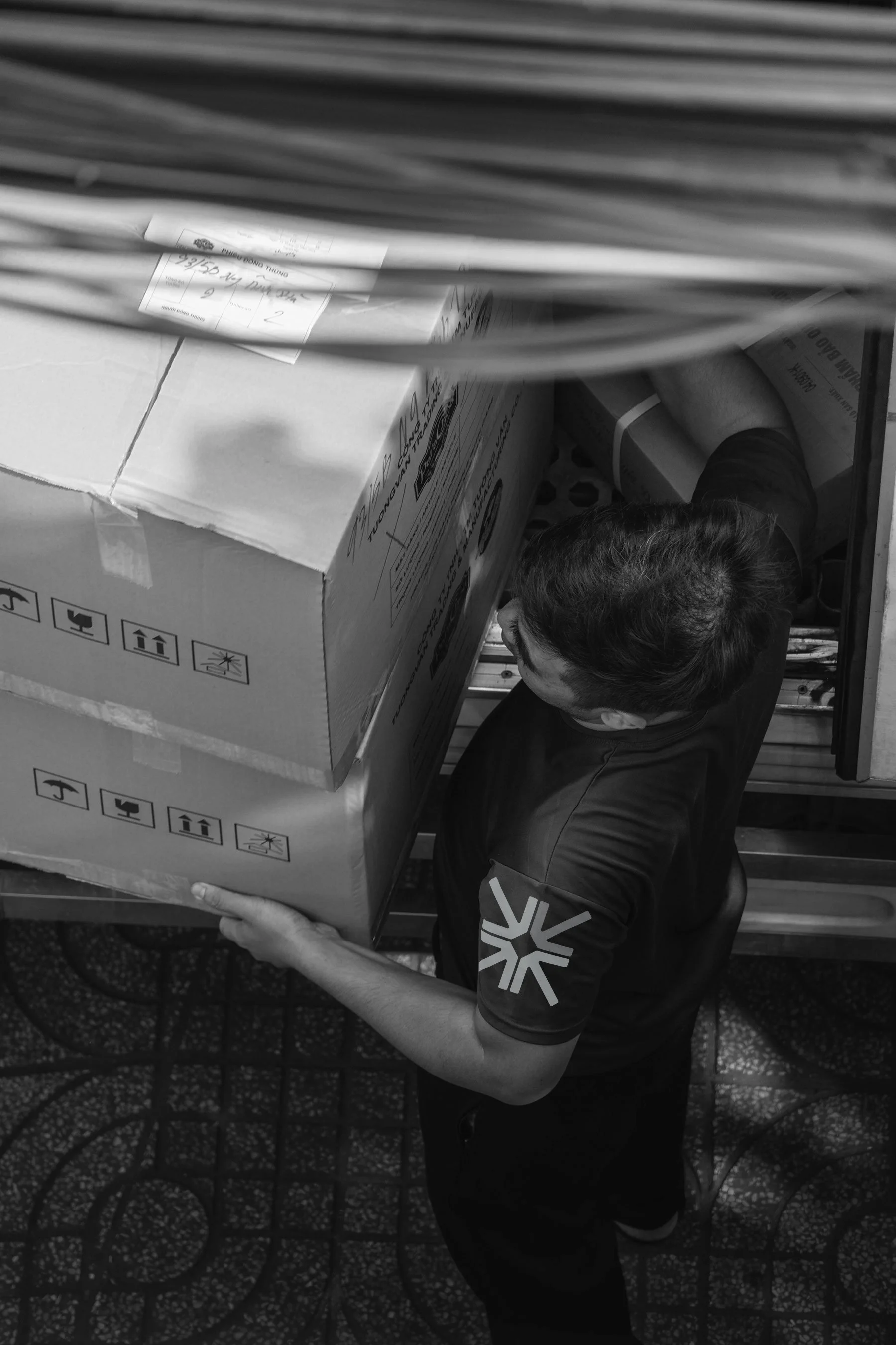 A person holding a large cardboard box on a patterned tile floor, with other boxes and packages visible around.