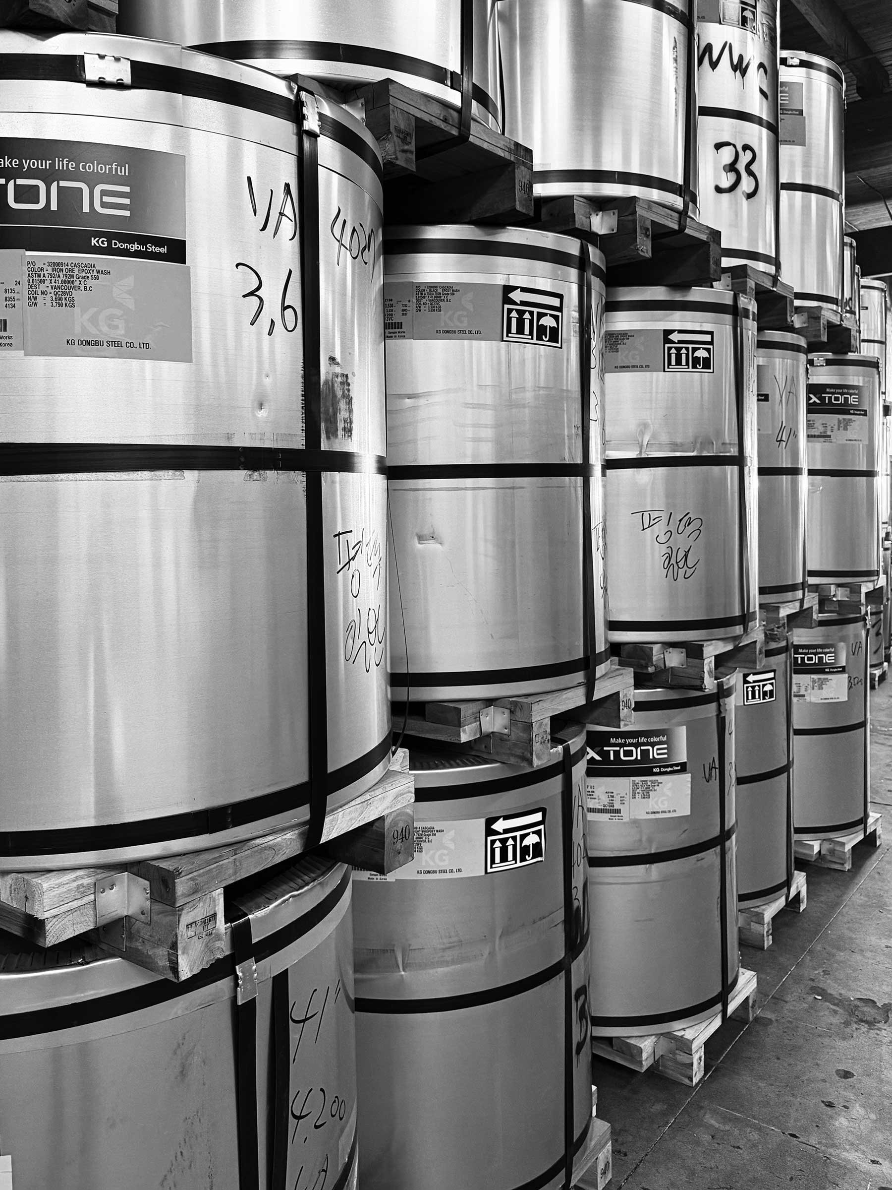 Stacked large metal drums on wooden pallets in a warehouse, some with handwritten labels and shipping symbols.