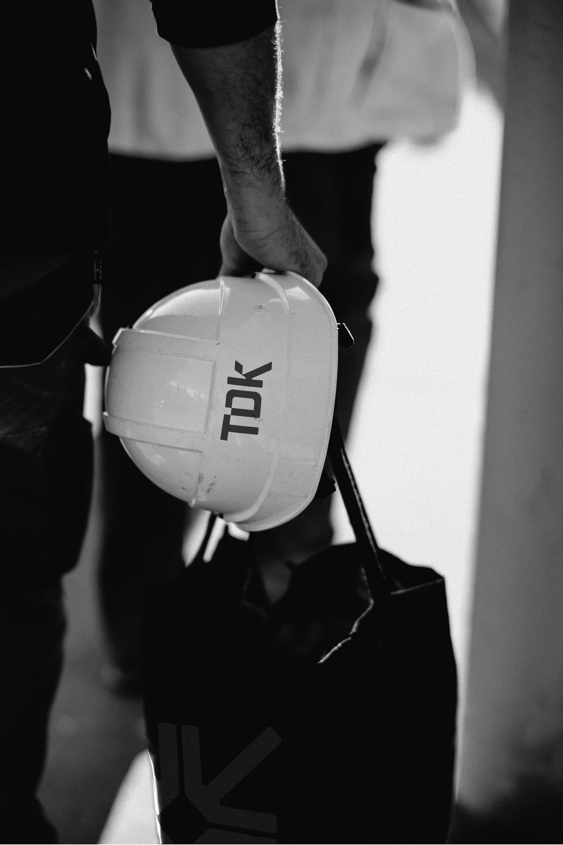 Close-up of a person carrying a white hard hat with the brand name TDK in black, wearing dark clothing with part of a logo visible on the pants, silhouette style in black and white.