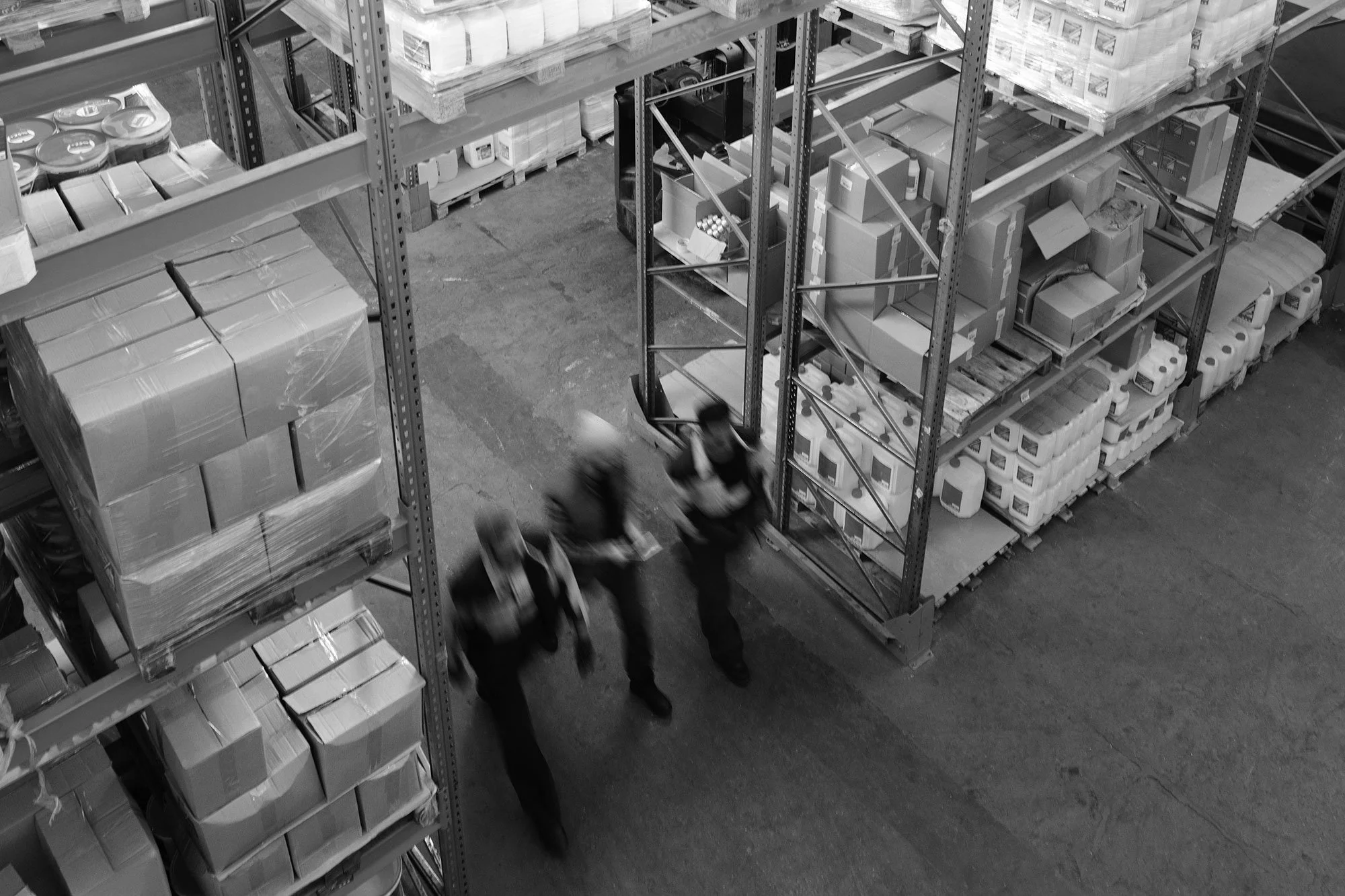 Black and white photo of a warehouse aisle with metal shelving loaded with boxes and bottles, and two blurred workers walking.