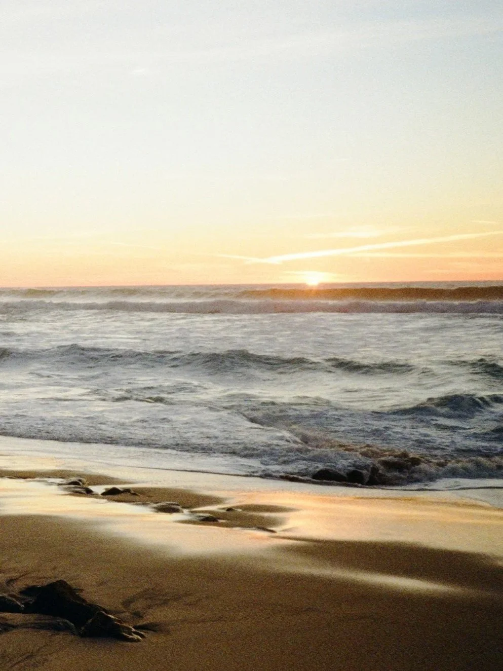 Sunset over the ocean with waves crashing on the sandy beach, some rocks and driftwood in the foreground.