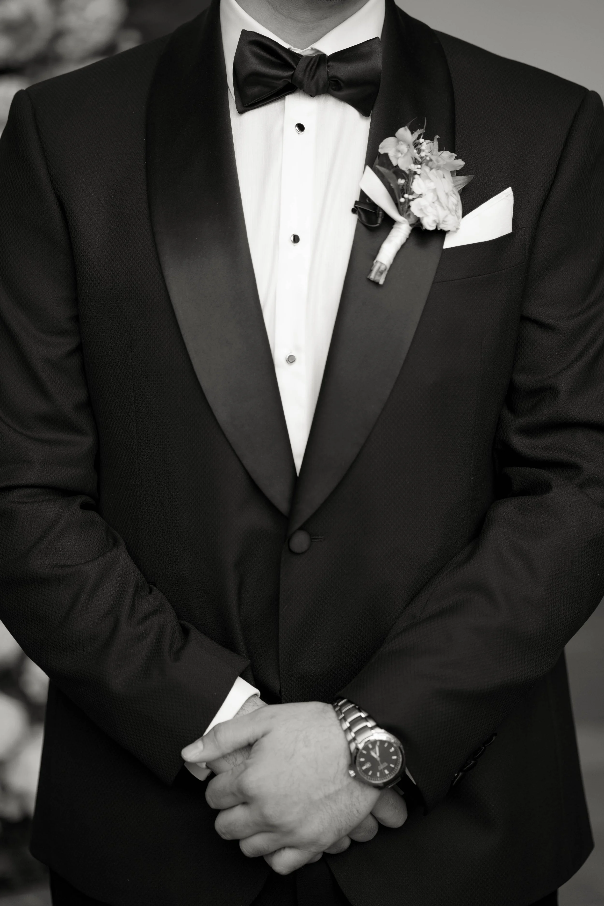 Close-up of a man in a black tuxedo with a white shirt, black bow tie, cufflinks, a wristwatch, and a boutonniere, with his hands crossed in front.